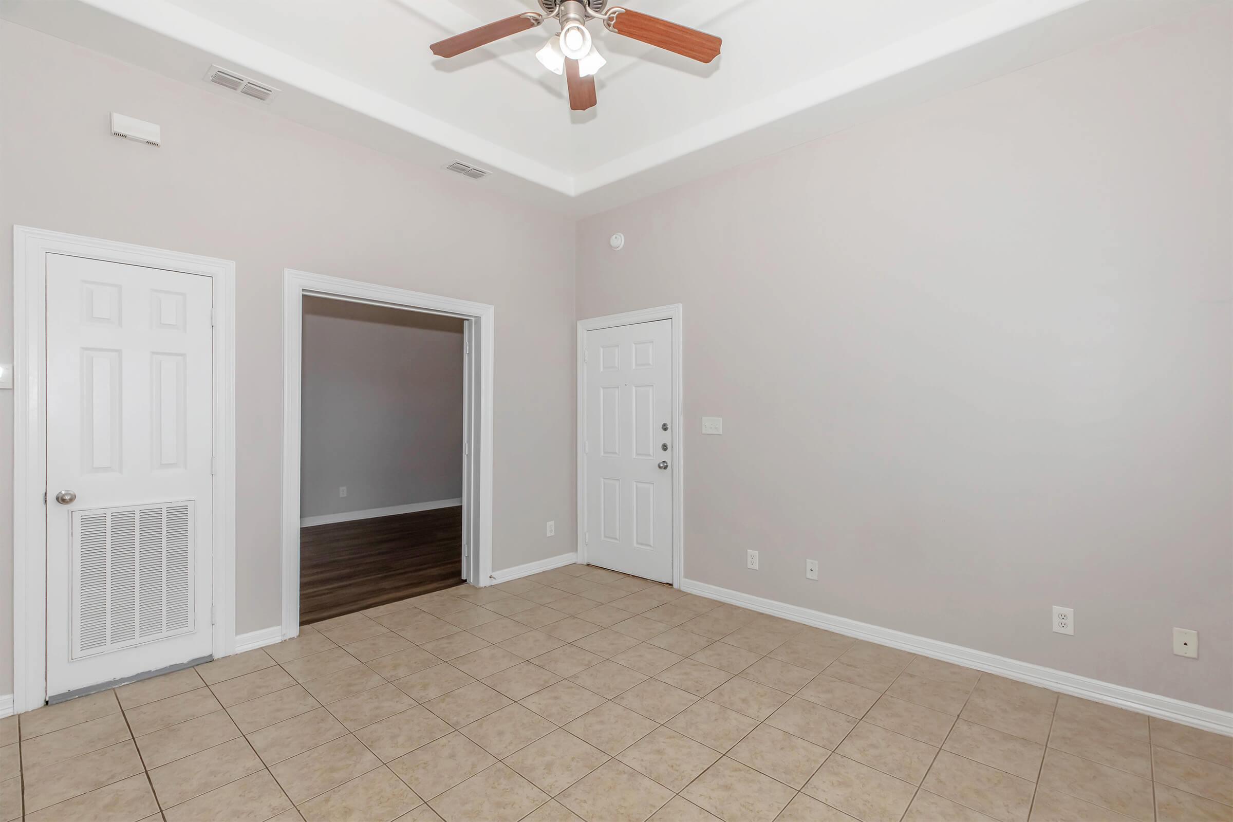 A spacious room with light beige walls and tiled flooring. There are two white doors on the left, one leading to an adjoining room with dark wooden flooring. The room features a ceiling fan with wooden blades and a white door entrance on the right. Natural light brightens the space.