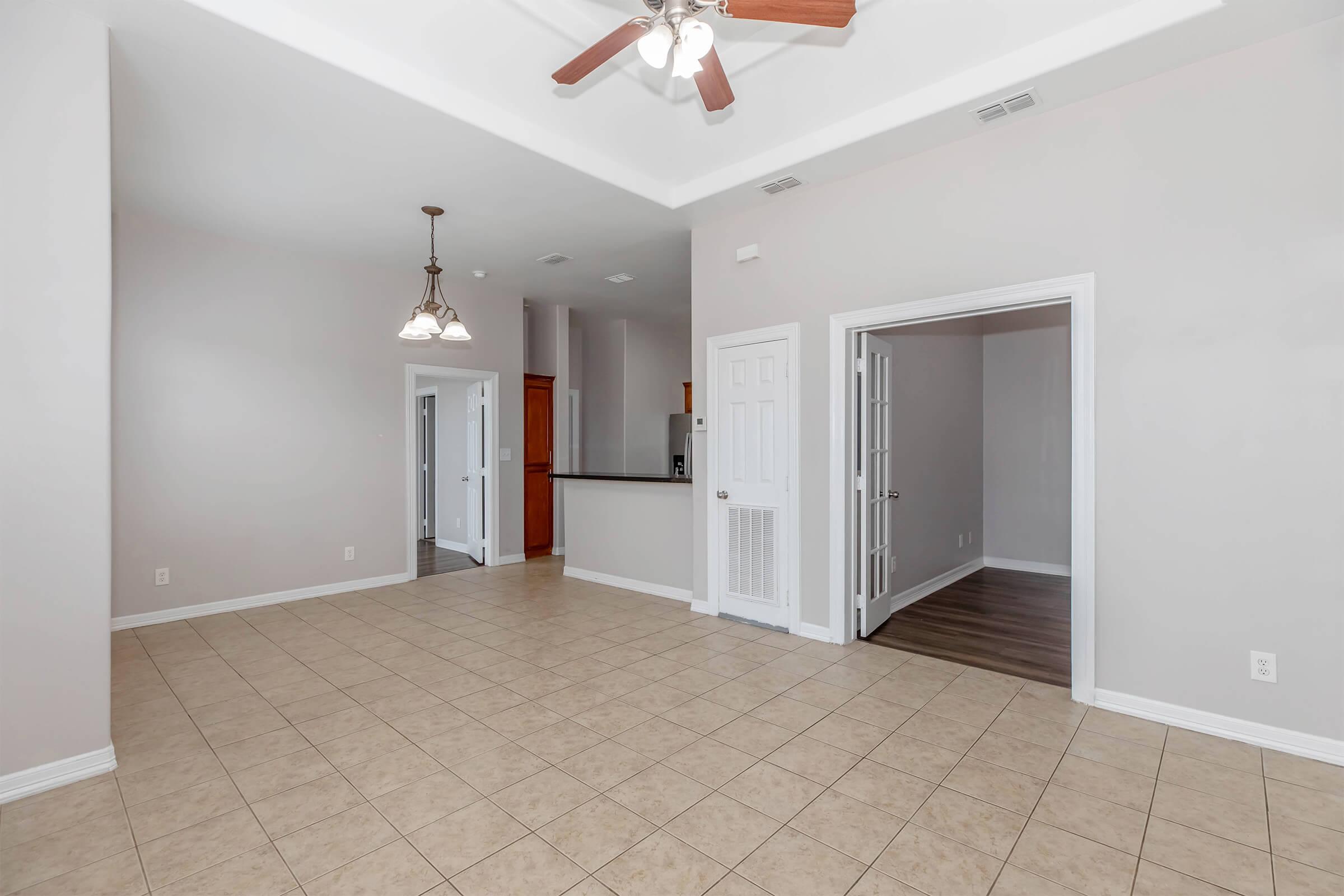 View of a spacious, empty room with beige tile flooring, features a ceiling fan, and two doorways leading to other rooms. There are light fixtures hanging from the ceiling, and the walls are painted in a light color. A kitchen area is visible in the background through an open doorway.