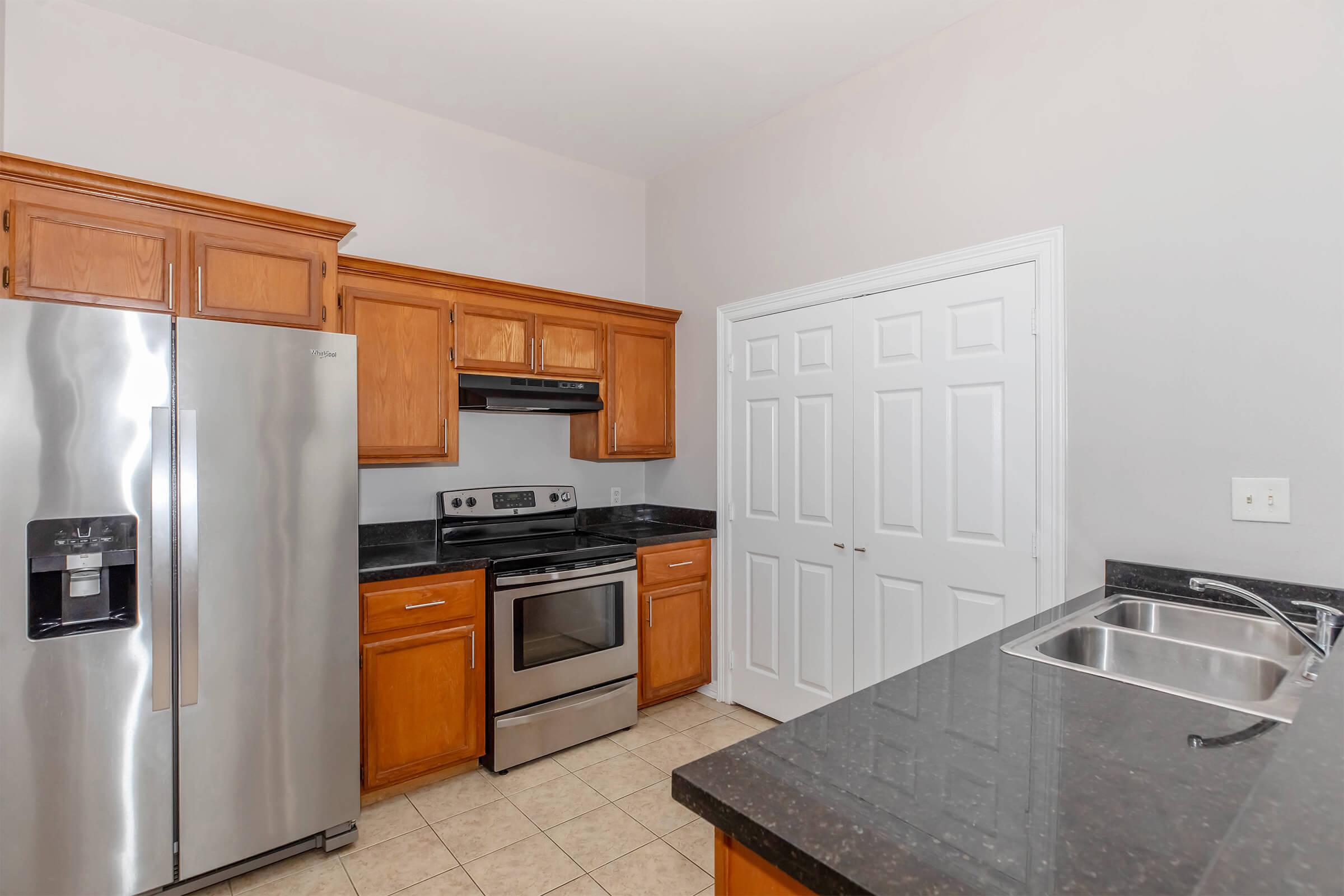 A modern kitchen featuring stainless steel appliances, including a refrigerator and oven, surrounded by wooden cabinetry and granite countertops. The kitchen has tile flooring and a double sink, with a doorway leading to another room in the background.