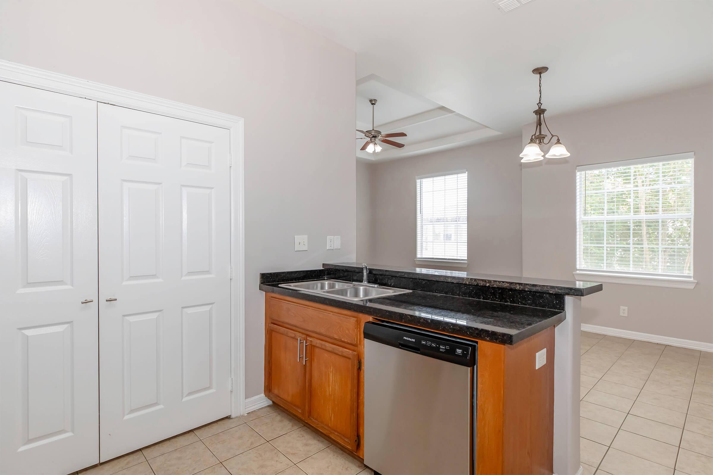 A clean, modern kitchen featuring a granite countertop, stainless steel dishwasher, and double sink. There are white cabinets and a light fixture above the island. The walls are painted light gray, and a window allows natural light to brighten the space.