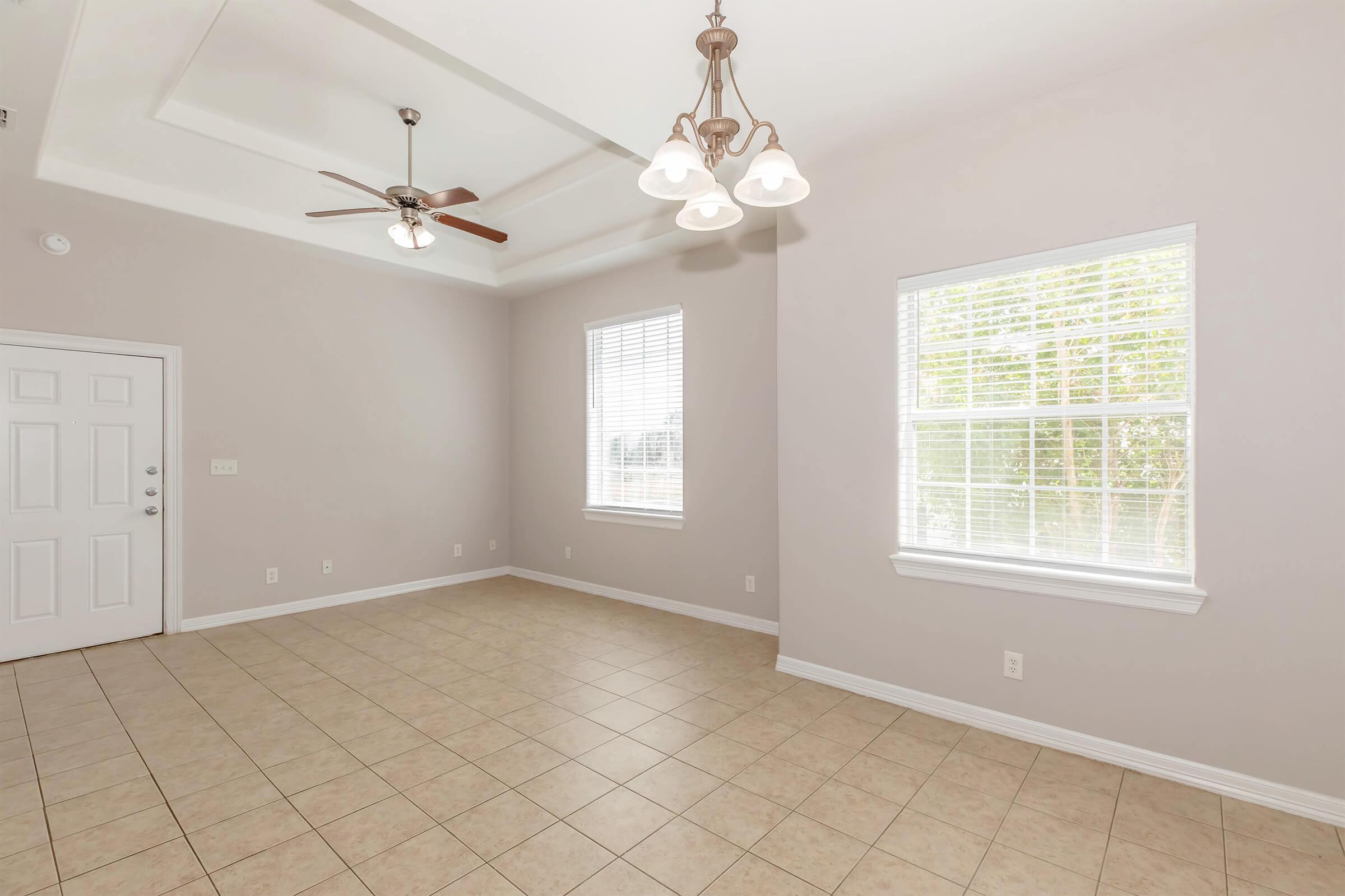 Spacious room featuring light-colored walls and tiled flooring. There is a ceiling fan with three lights and large windows allowing natural light. The room is empty, offering a versatile space for various furniture arrangements.