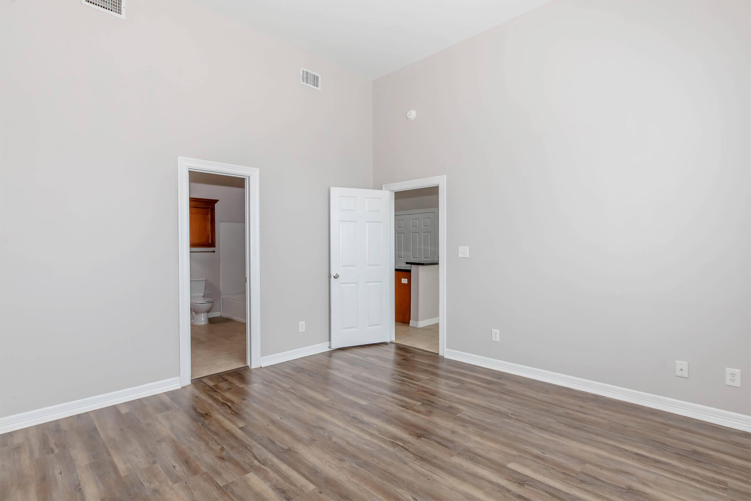 A spacious, empty room with light gray walls and laminate flooring. A doorway leading to a bathroom with visible fixtures and another doorway leading to a kitchen area is in the background. The room is well-lit with natural light, creating a bright and airy atmosphere.