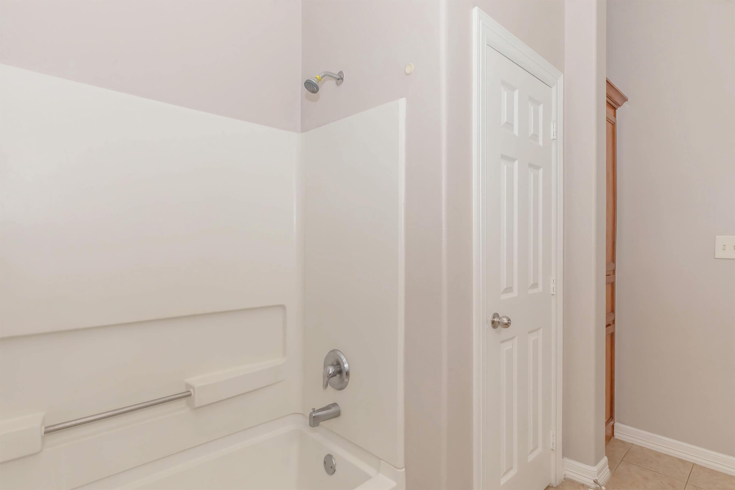 A clean, modern bathroom featuring a white bathtub with a showerhead attached. The walls are painted a light color, and there is a closed white door leading to another room. The floor is tiled, and a light fixture is visible on the wall.