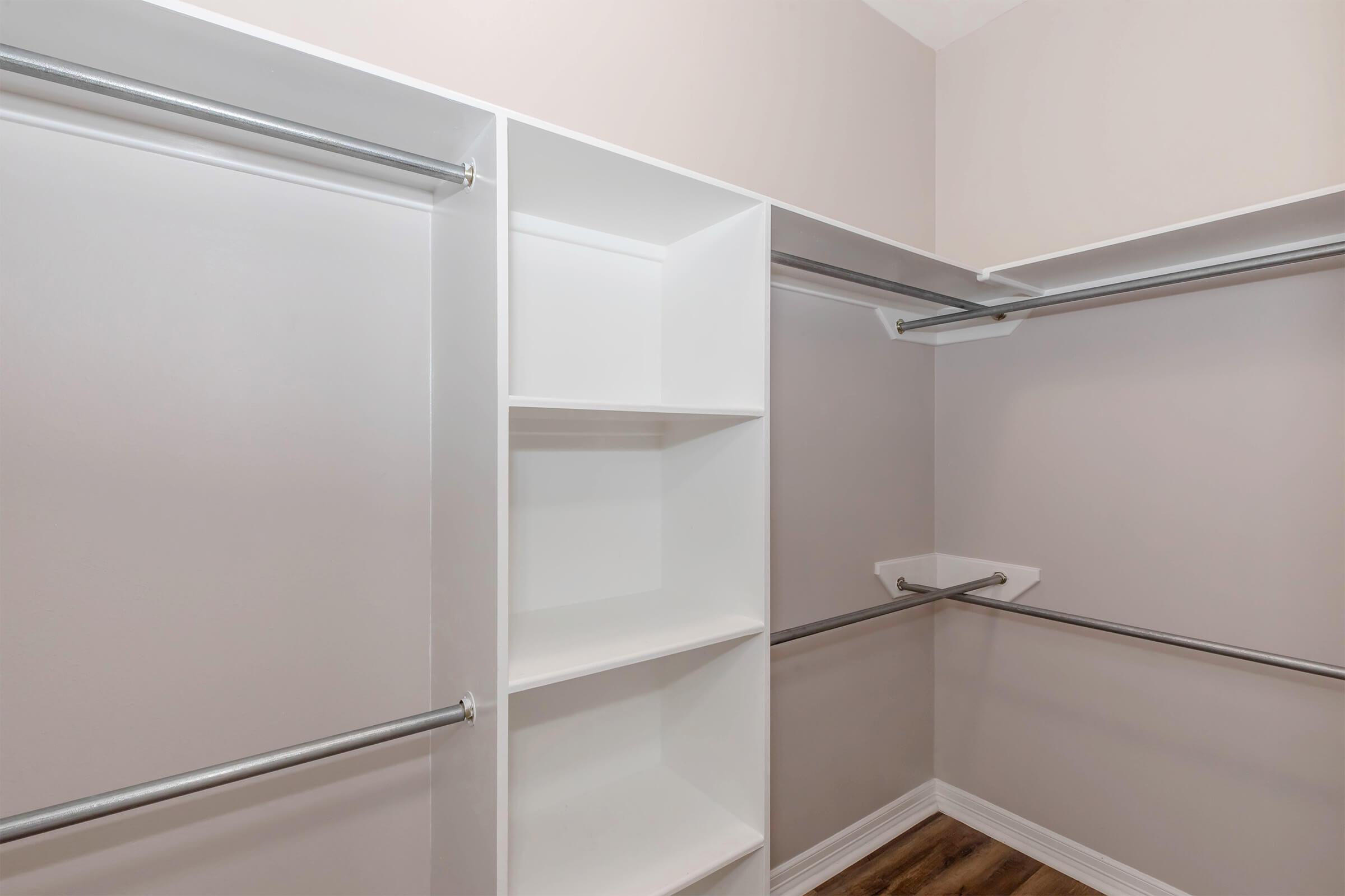 Spacious walk-in closet featuring white shelving units and two hanging rods against a soft gray wall. The floor is made of wood, adding warmth to the minimalist design. The closet is empty, showcasing ample storage space for clothes and accessories.