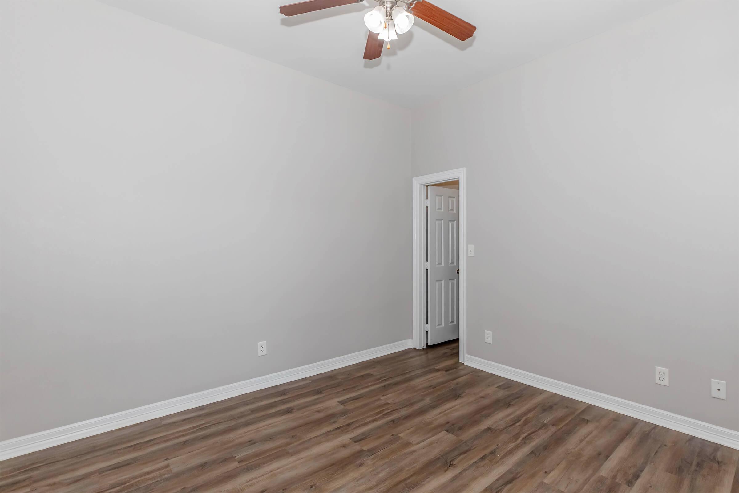 Empty room with light gray walls and a ceiling fan. The floor is covered in brown laminate. A door is visible on the right side, leading to another space. The room has a clean and simple aesthetic, with no furniture or decorations present.