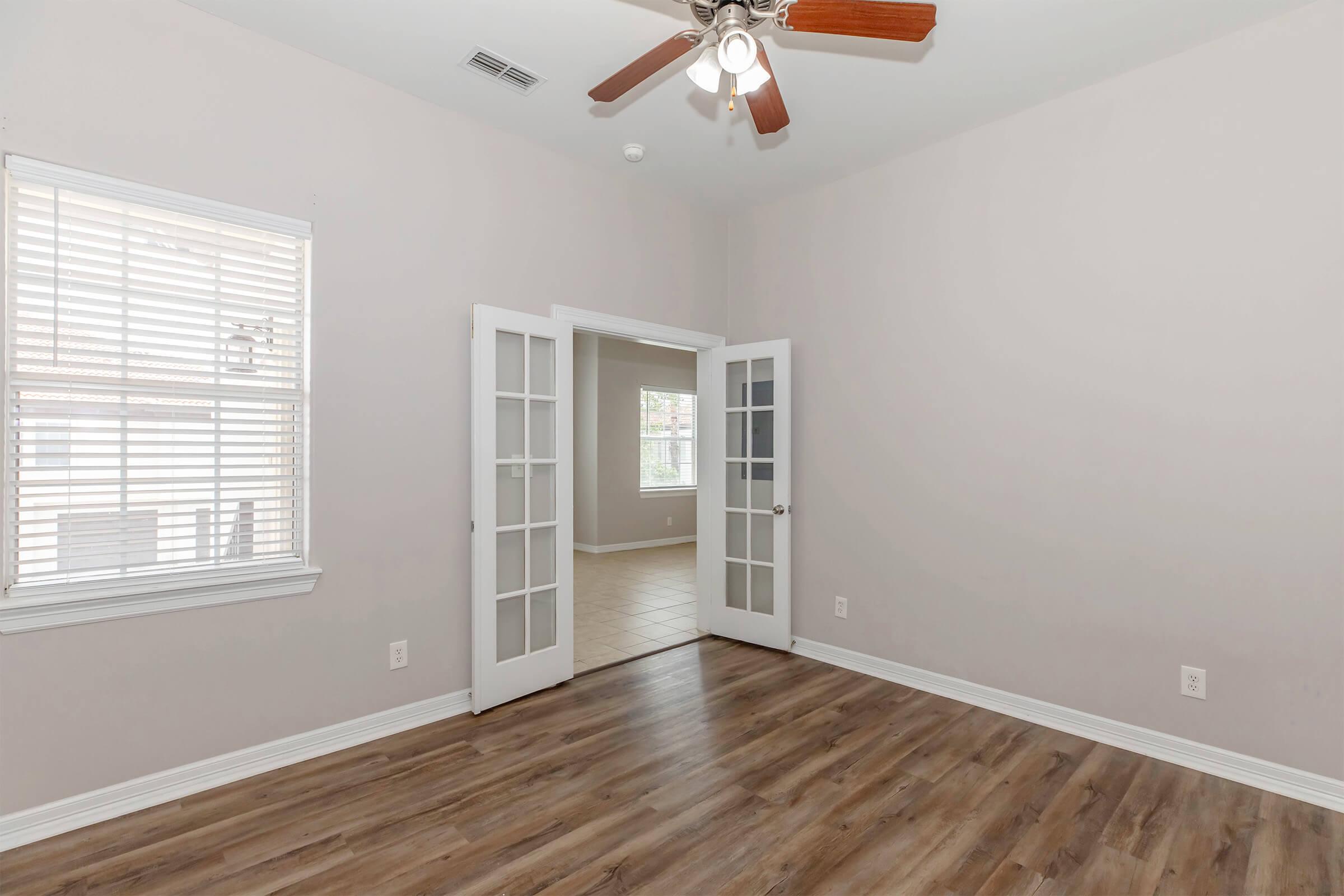 A bright, unfurnished room with light gray walls and hardwood flooring. Two sets of double doors with glass panels open into the space, and a ceiling fan is installed above. Natural light enters through a window with white blinds, illuminating the room's simplicity and potential.