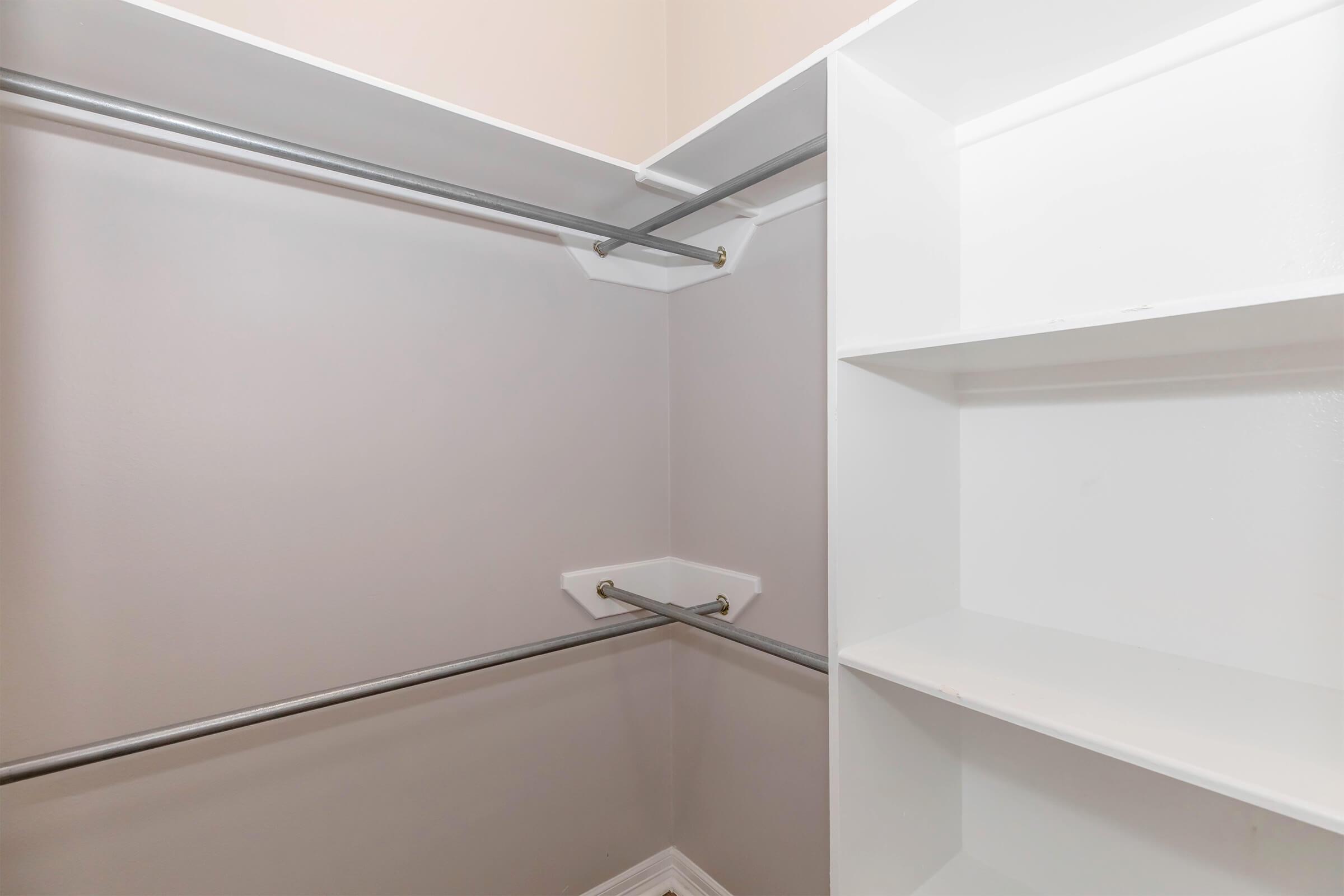 A spacious, empty closet with light gray walls. Two metal hanging rods are installed along one wall, and there are white shelf units on the adjacent wall, creating organized storage space. The overall design is clean and minimalistic.