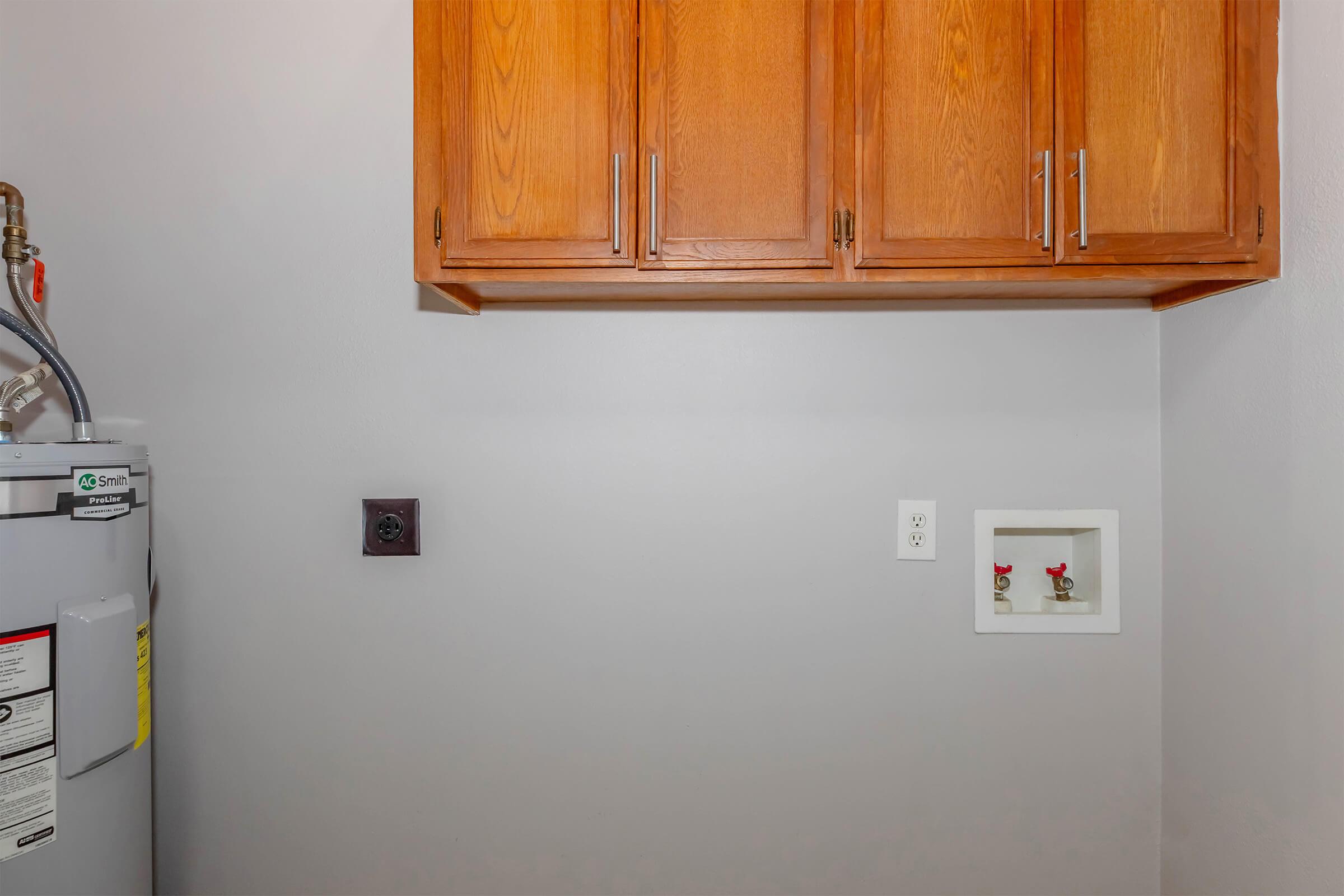 A utility area featuring several wooden cabinets above a water heater. The walls are painted light gray. On the right side, there are electrical outlets and a small white box with two red valves visible. The overall space is tidy and functional, typically used for laundry or storage purposes.