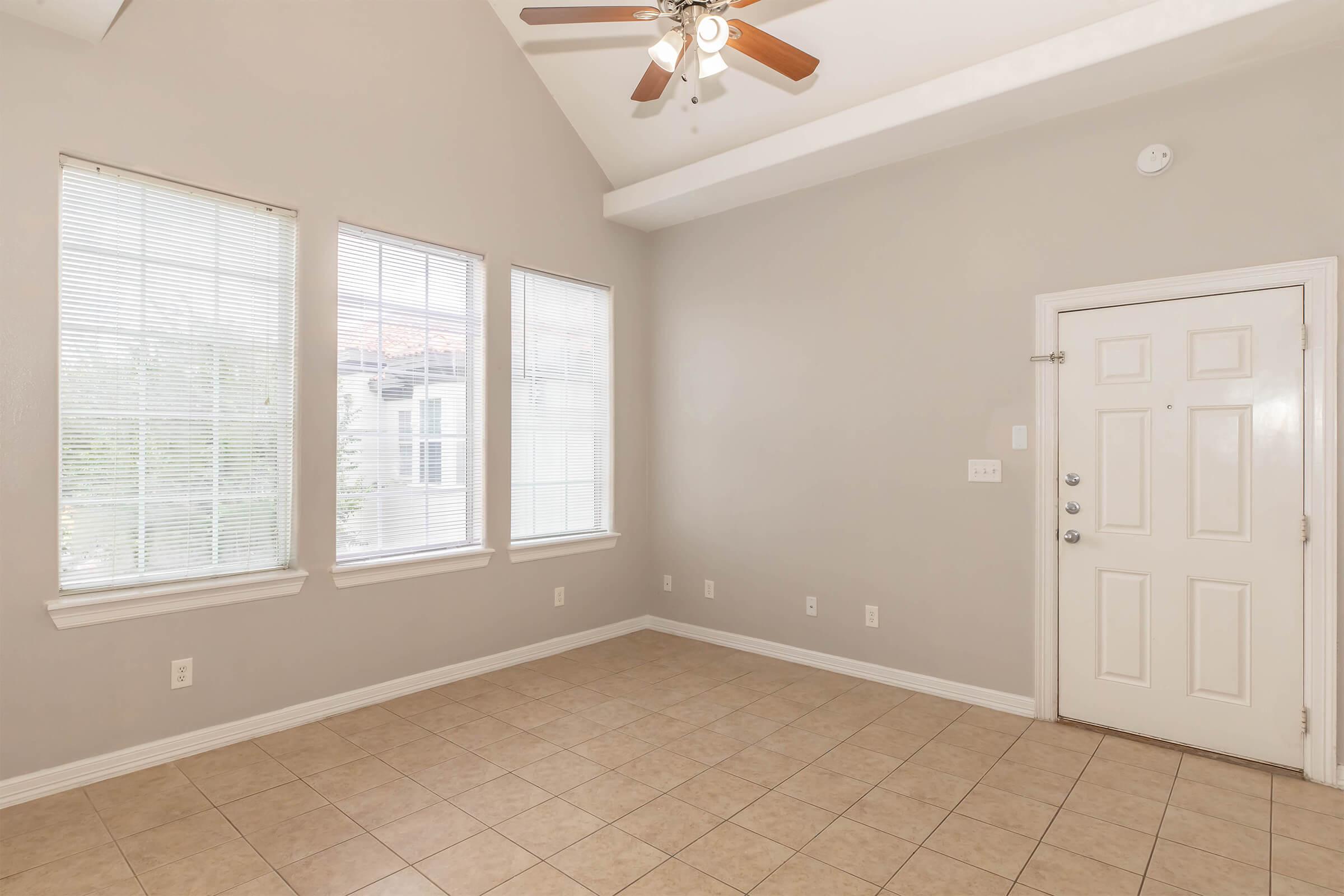 A spacious, empty room with light-colored walls and large windows that allow natural light. The room features a ceiling fan and tile flooring, with a door leading outside visible on the right side. The overall atmosphere is bright and airy, ideal for various potential uses.