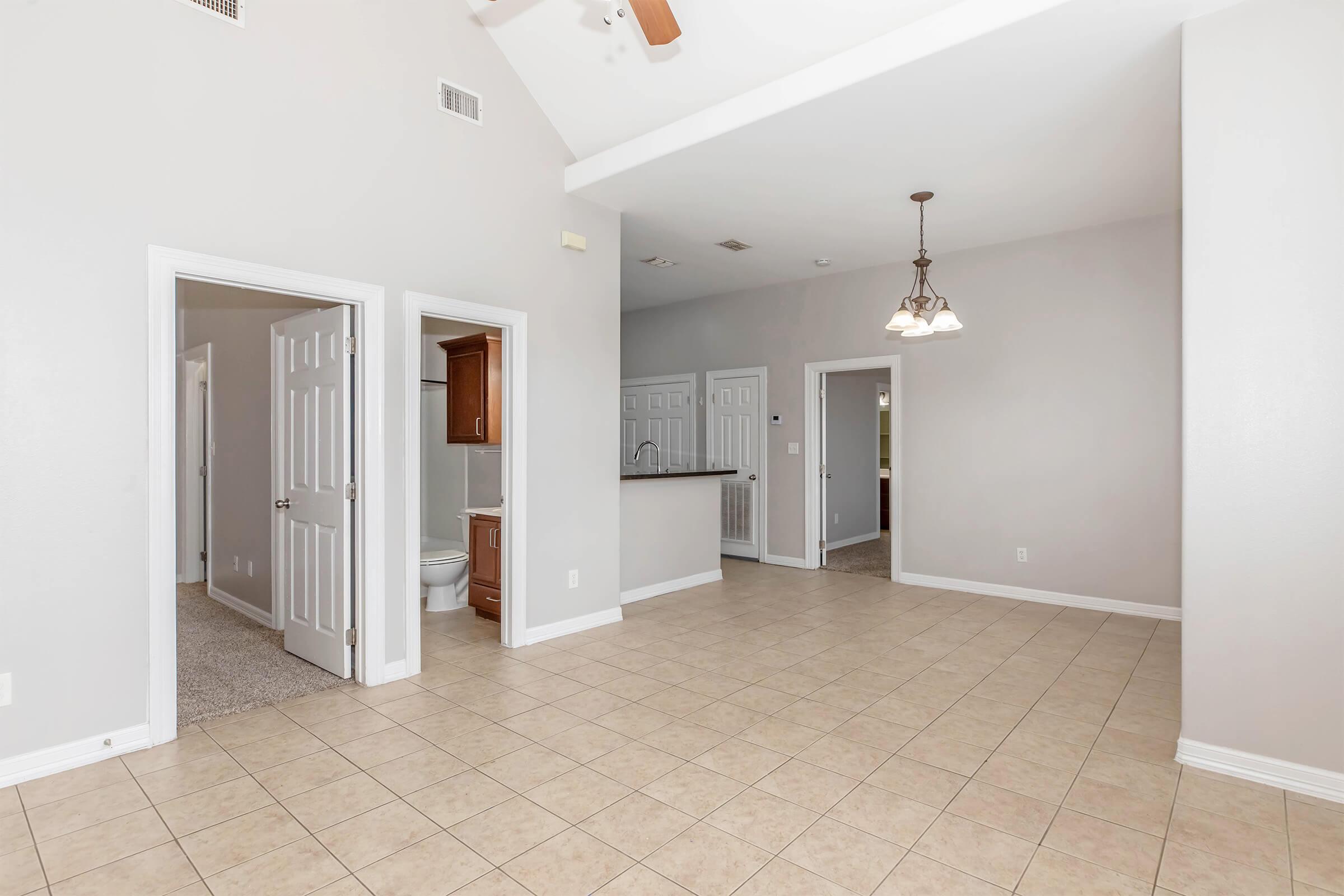 Spacious interior of a home featuring tiled flooring, a high ceiling, and neutral-colored walls. The image shows multiple doorways leading to different rooms, with one door opening to a bathroom. A light fixture hangs from the ceiling, enhancing the bright atmosphere.