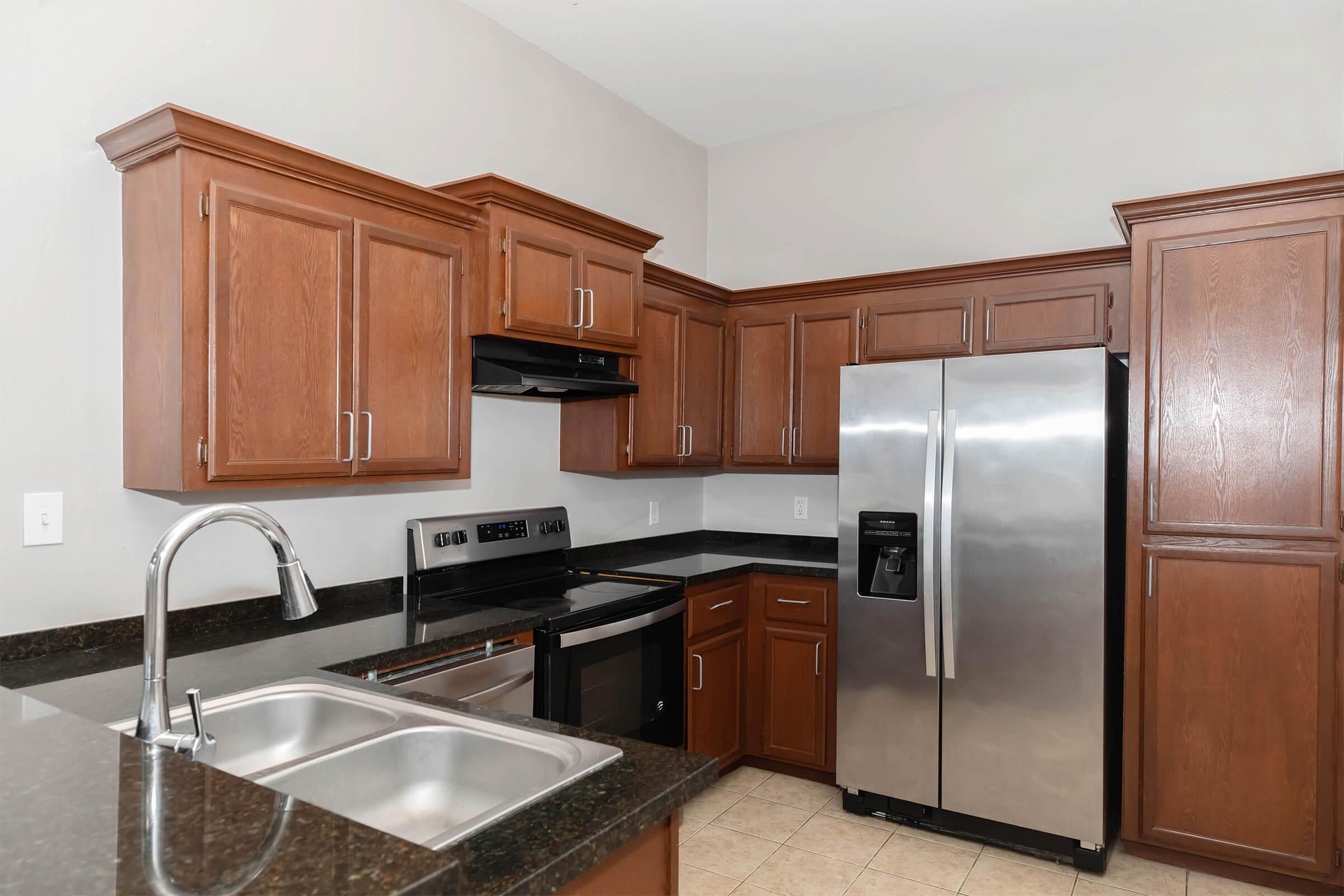 Modern kitchen featuring brown wooden cabinets, granite countertop, stainless steel refrigerator and stove, and a double sink. The walls are painted light gray, and the tile floor adds a touch of elegance.