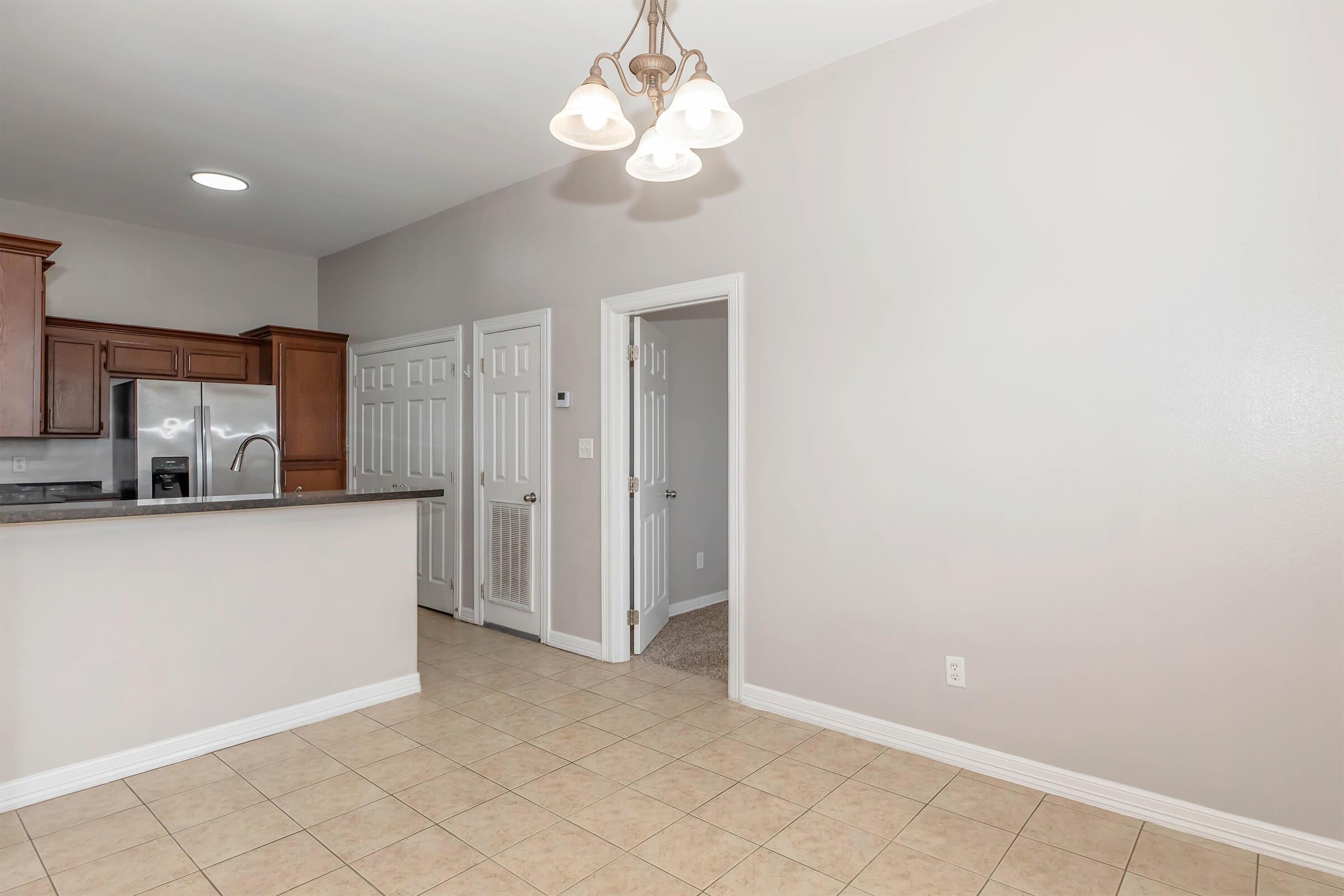 Interior view of a modern kitchen showcasing wooden cabinetry, stainless steel appliances, and a tiled floor. A doorway leads to additional rooms, with light-colored walls and a ceiling light fixture adding brightness to the space. The layout appears open and inviting.