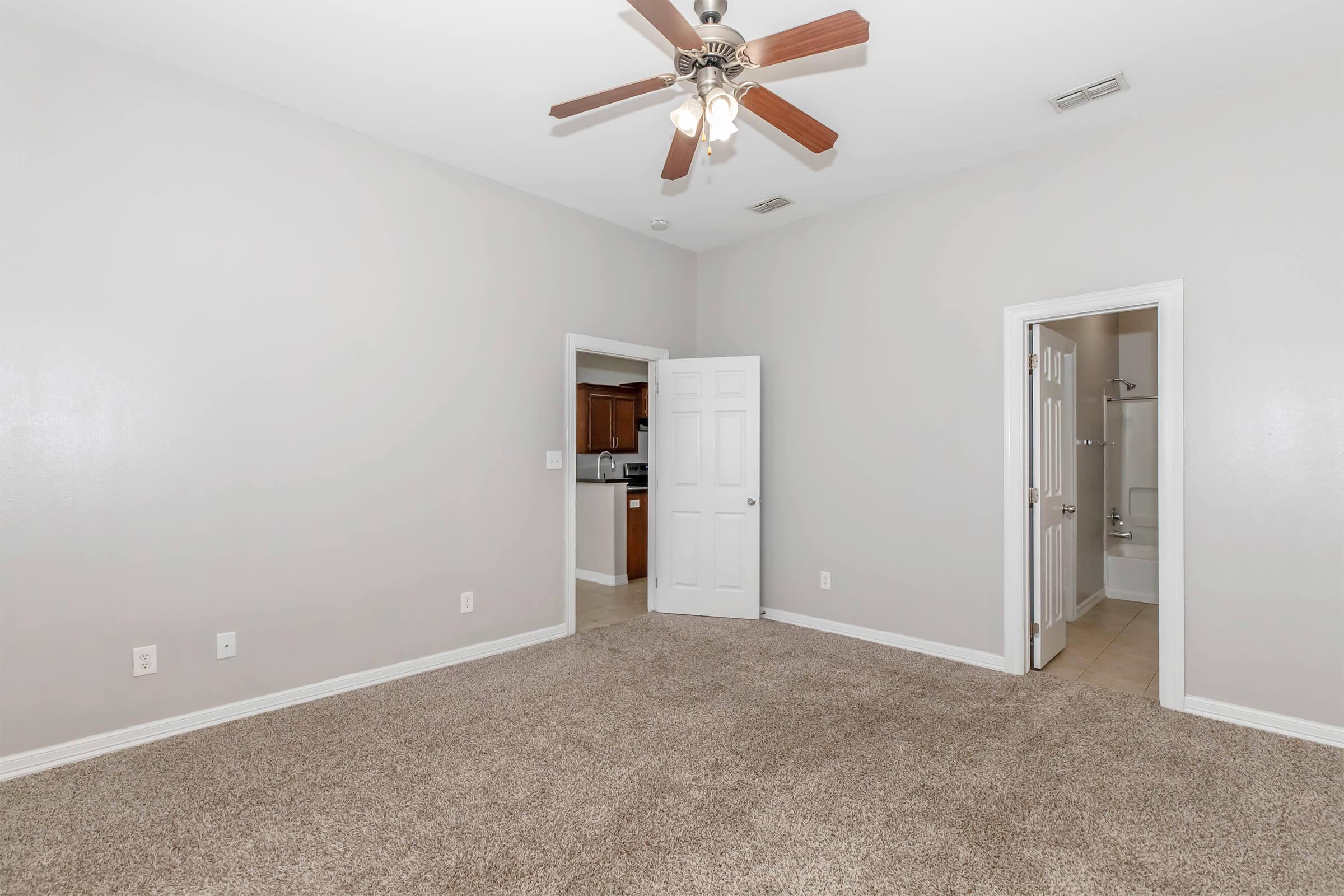 A spacious bedroom featuring light gray walls, a ceiling fan, and plush carpet. To the left, a doorway leads to a kitchen area with wooden cabinets. A bathroom is visible through another door. Natural light enters, creating a bright and airy atmosphere.