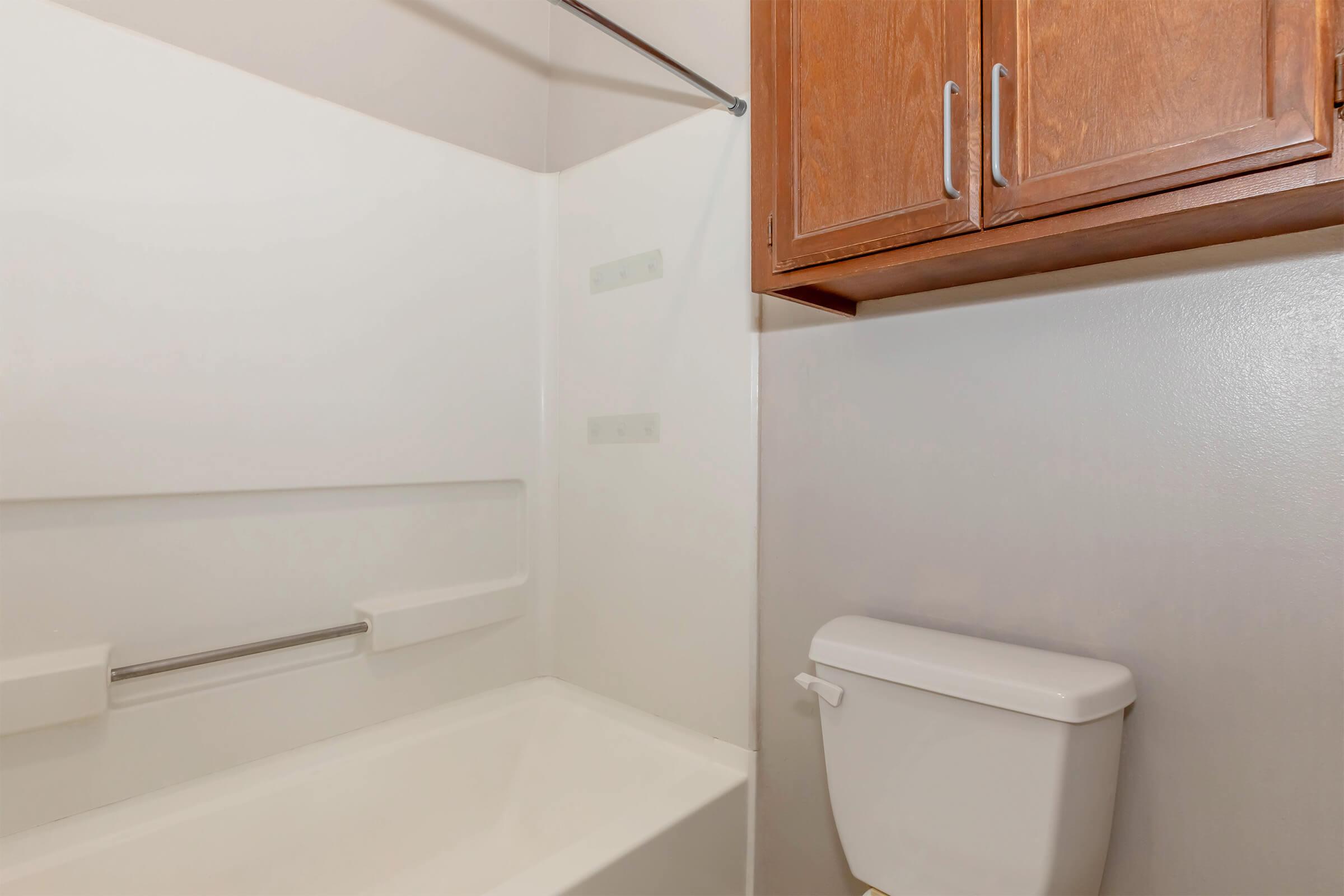 A clean bathroom with a white bathtub, a wall-mounted support rail, and a toilet. Above the toilet, there are wooden cabinets for storage. The walls are painted a light color, creating a bright and airy atmosphere.