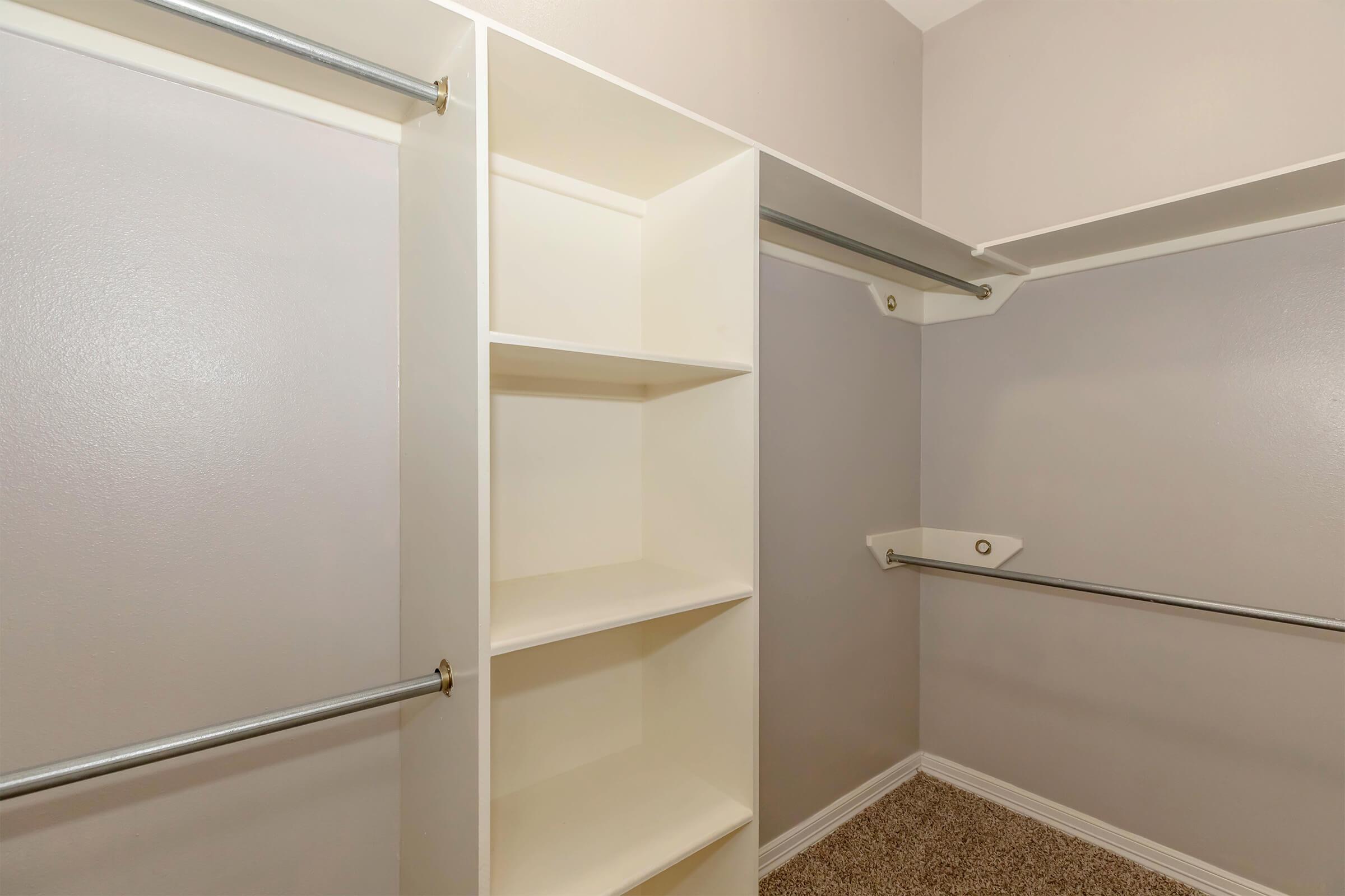 A well-organized closet with light-colored walls, featuring multiple hanging rods and a built-in shelving unit with open compartments, on a plush carpeted floor.
