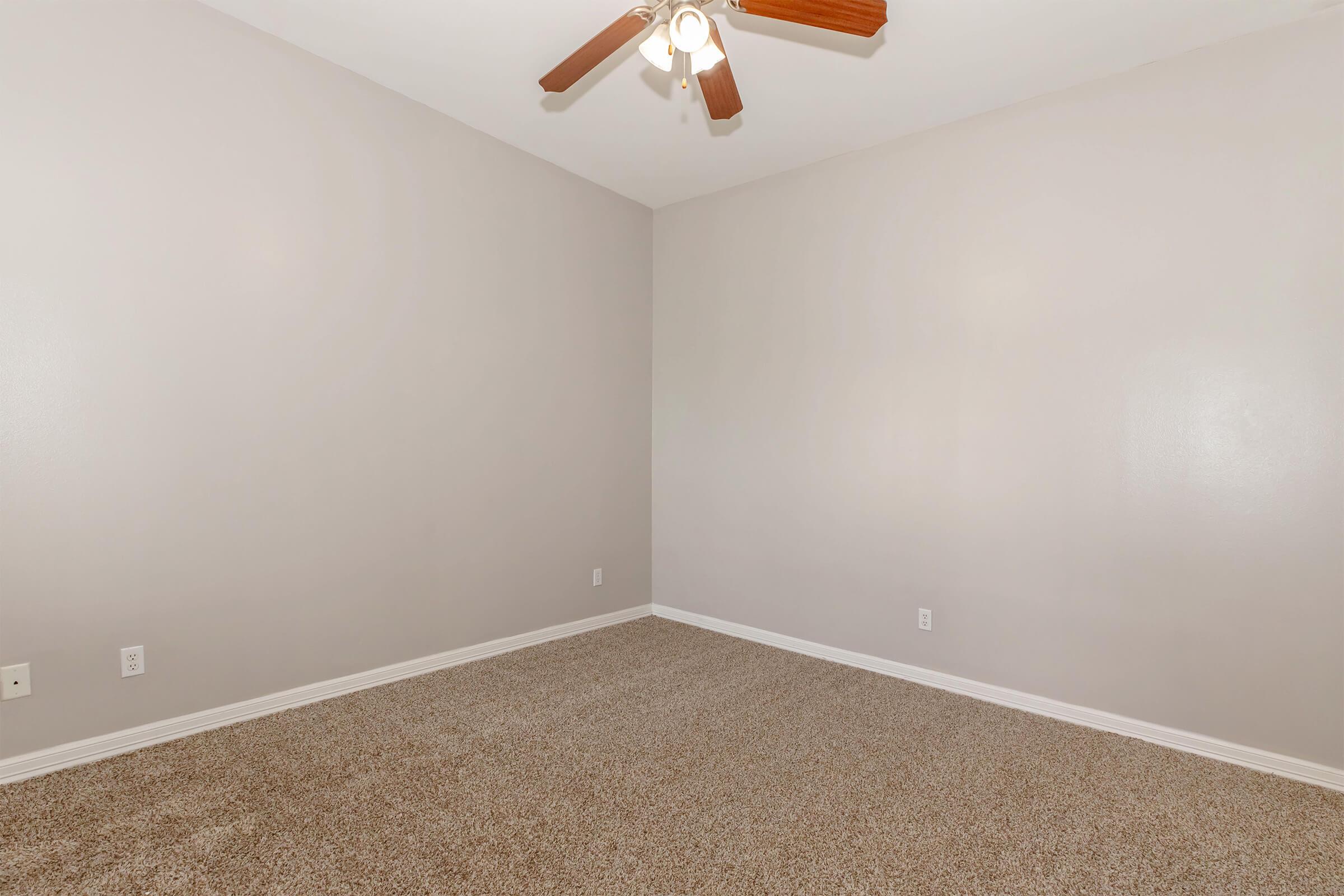 Empty room with light gray walls, a carpeted floor, and a ceiling fan. The room has no furniture or decorations, creating a spacious and minimalistic appearance. Natural light may be entering from an unseen window.