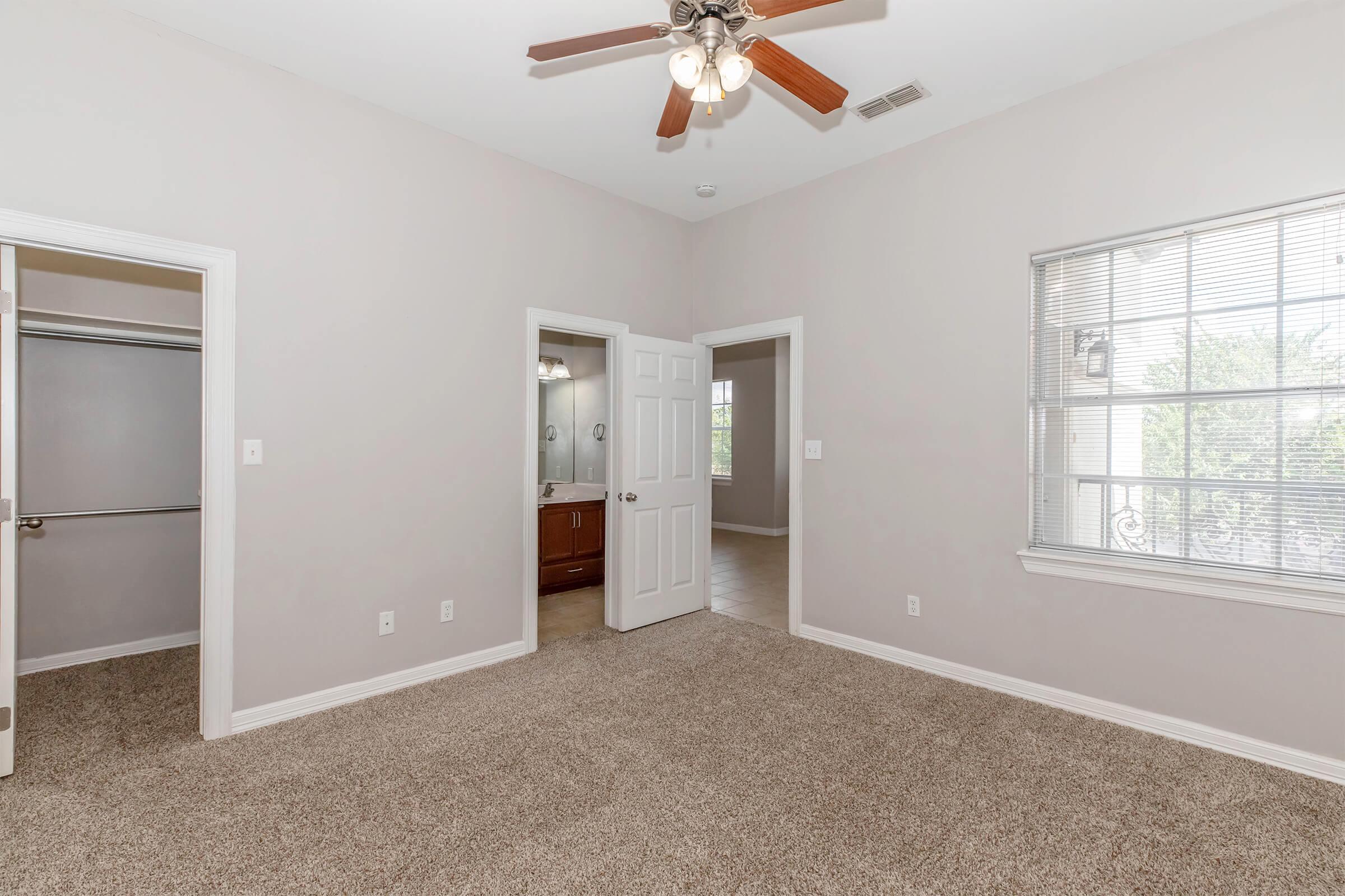 Spacious bedroom with light-colored walls and carpet flooring. Features a ceiling fan, a window with blinds allowing natural light, and a doorway leading to a bathroom with a vanity. A closet can be seen through an open door on the left. Overall, the room has a bright and airy feel.