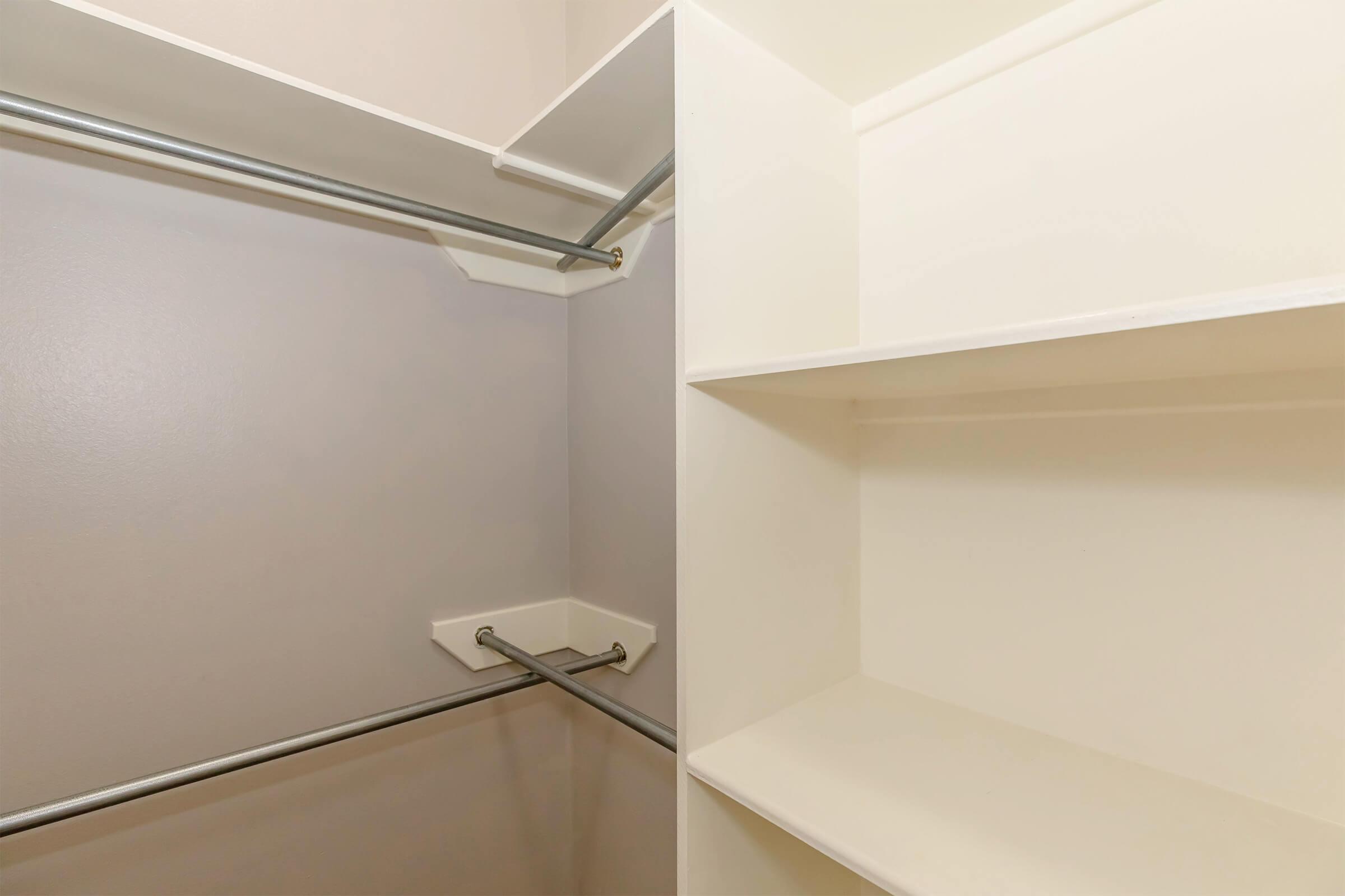 Empty closet with white shelving and two horizontal metal rods for hanging clothes. The walls feature a light color, providing a clean and spacious appearance.