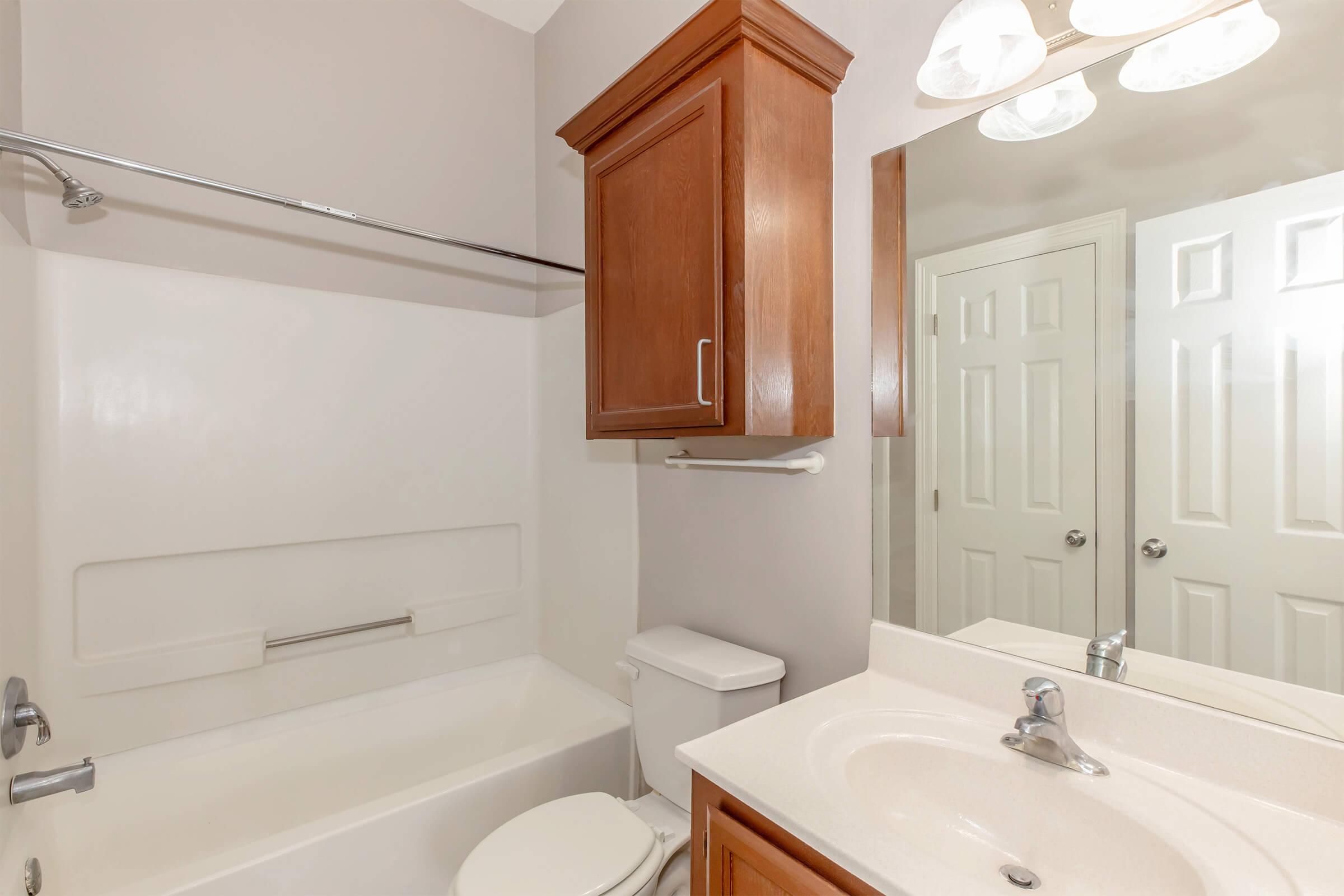 A clean and modern bathroom featuring a bathtub, toilet, sink with a countertop, a mirrored cabinet above the sink, and light fixtures. The walls are painted in a light gray color, and there is a towel bar beside the bathtub. The bathroom has a simple design and is well-lit.