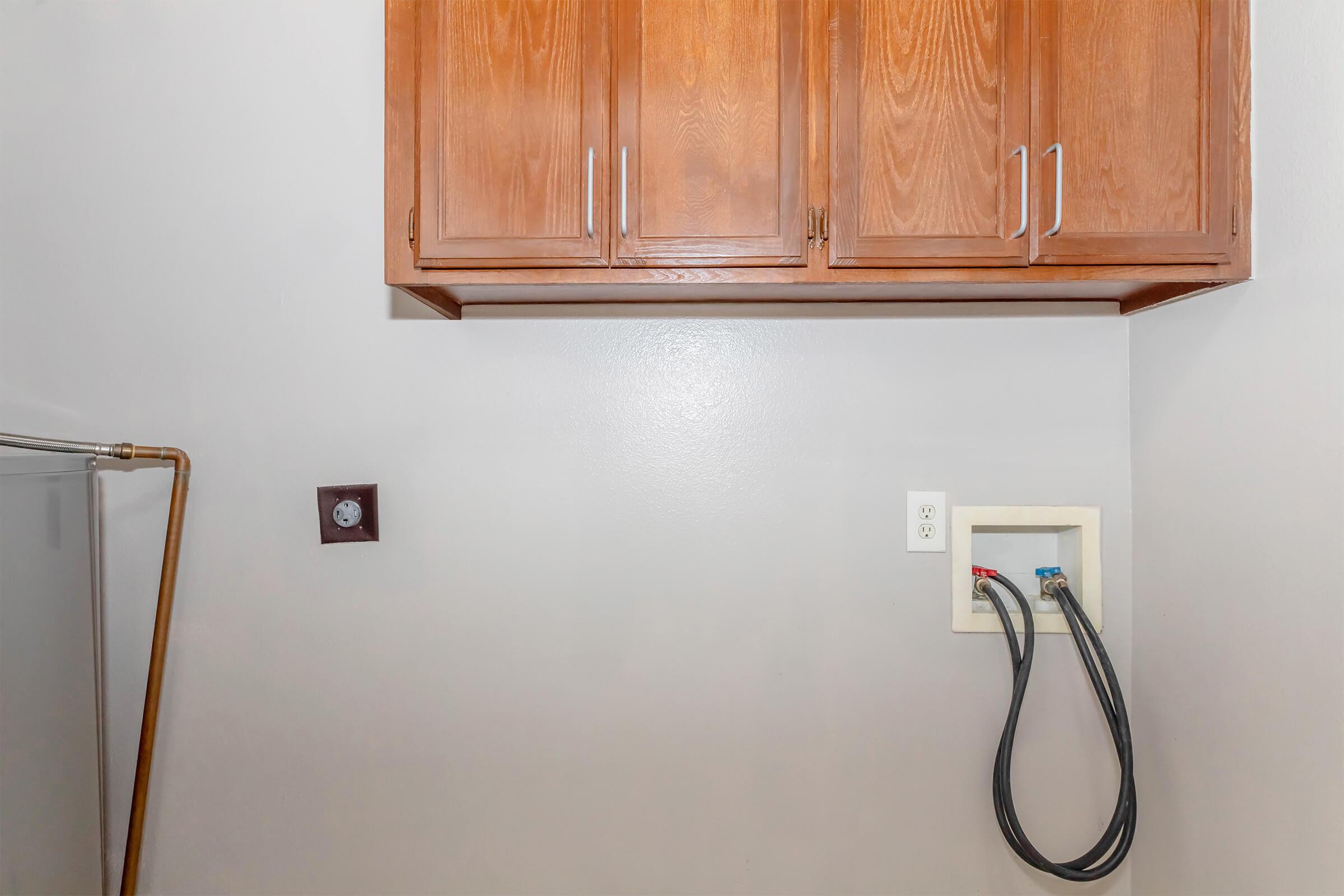A laundry room featuring wooden cabinets on the wall, a power outlet, and a wall-mounted connection for a washing machine. A portion of a shower curtain rod is visible in the corner, indicating possible versatility of the space. The walls are painted a light gray color.
