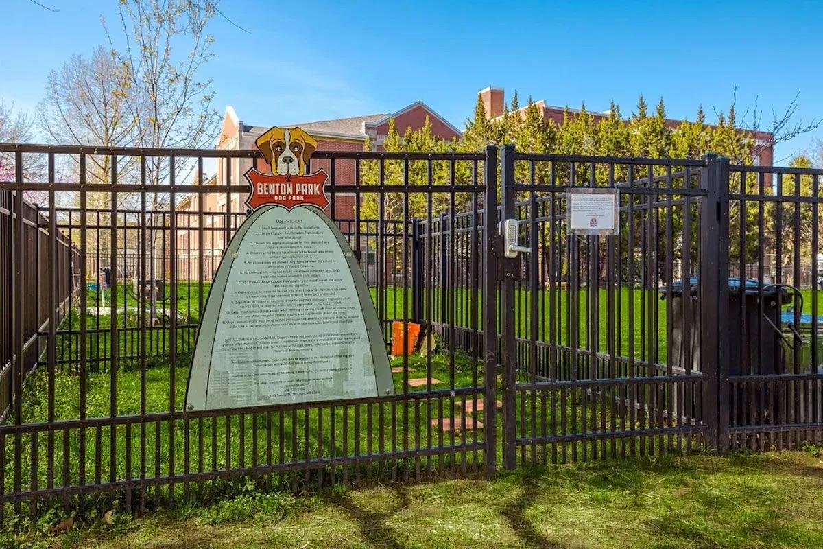 A fenced dog park area with a sign detailing park rules and regulations. Green grass is visible inside the park, along with a trash bin. In the background, trees and a building can be seen under a clear blue sky.