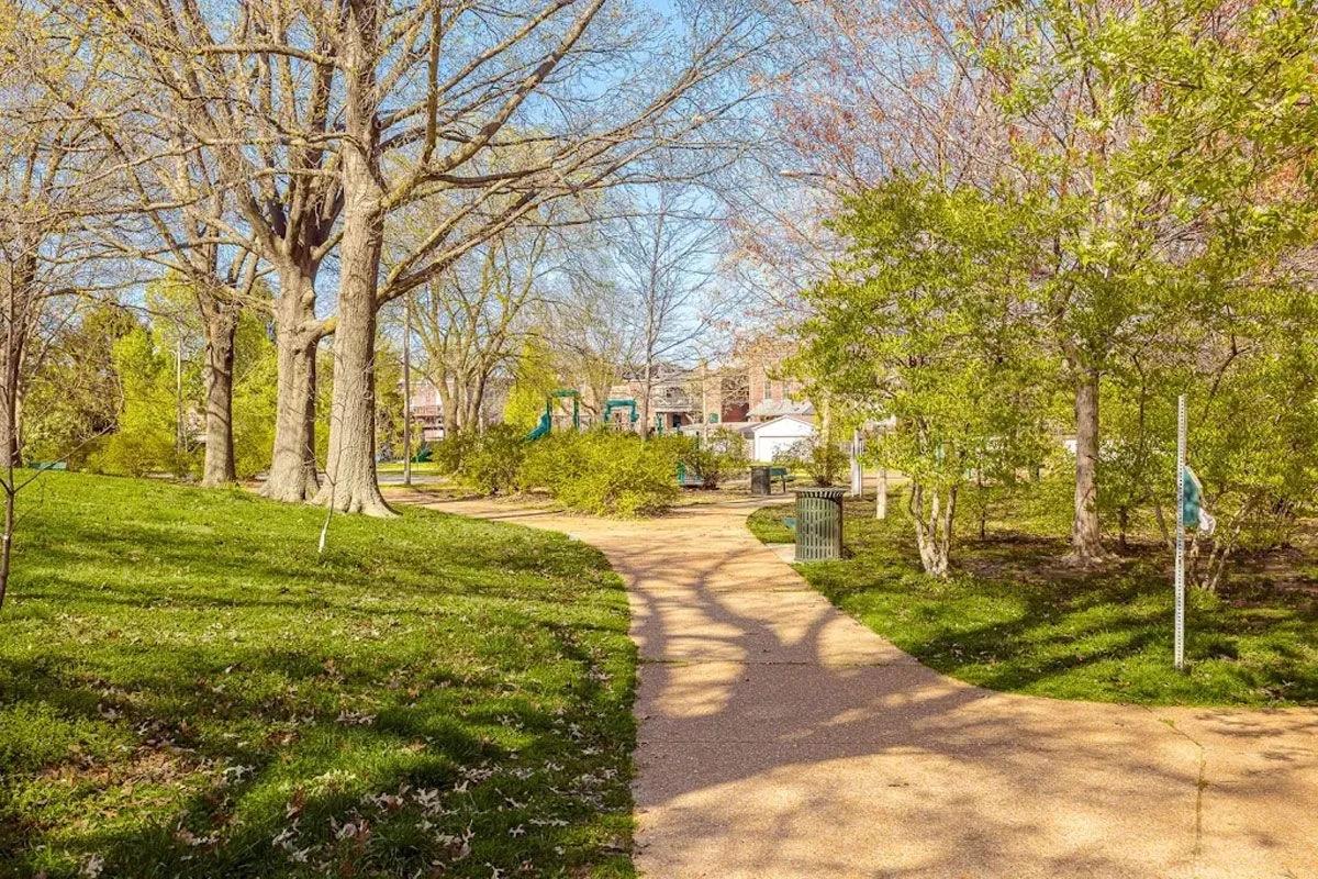 A serene pathway winding through a lush green park, bordered by tall trees with budding leaves. Sunlight filters through the branches, creating dappled shadows on the path. In the background, glimpses of playground equipment and benches can be seen, indicating a peaceful outdoor area for recreation.