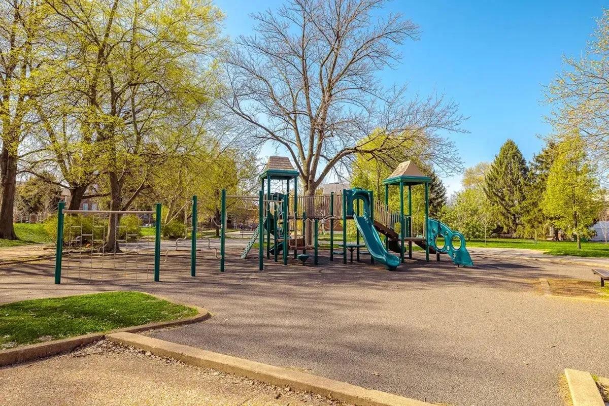 A playground in a park featuring green slides and climbing structures. Surrounding the playground are trees with fresh green leaves, and the ground is paved with a light-colored surface. The sky is clear and blue, creating a bright and inviting atmosphere.