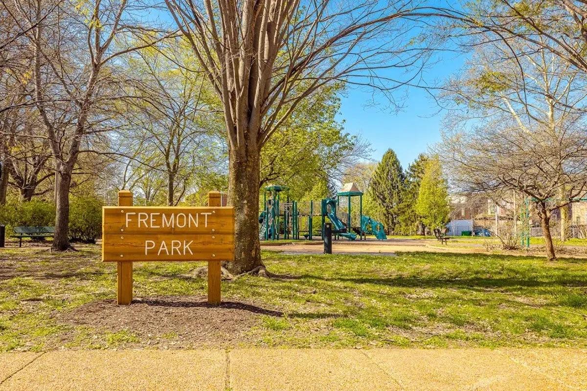 A wooden sign reading "Fremont Park" stands in a grassy area, with trees surrounding the park. In the background, a playground featuring green equipment is visible. The scene is bright and sunny, showcasing a serene outdoor space ideal for families and children.