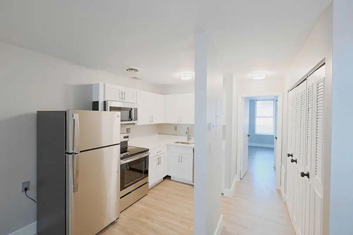 Modern kitchen with stainless steel refrigerator and stove, white cabinetry, and light-colored flooring. It features an open layout leading to a hallway with doors to closets. Bright lighting enhances the spacious feel of the area.
