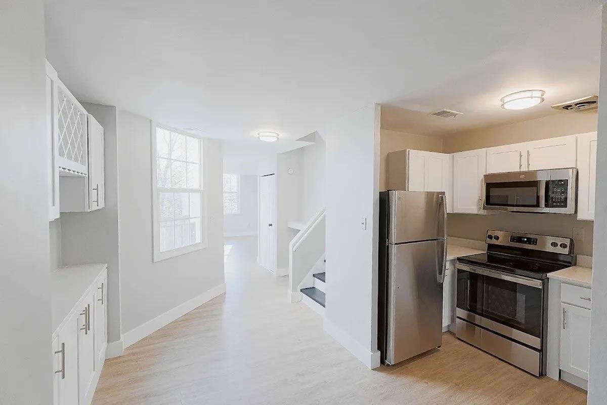 Bright kitchen area with white cabinets, stainless steel appliances, and a modern design. Features an open layout leading to a staircase, large windows allowing natural light, and clean, neutral-colored walls. A cozy and inviting atmosphere.