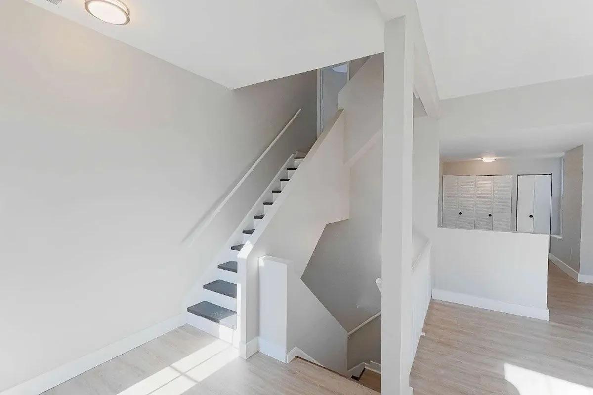A bright, modern interior view of a staircase leading to the upper floor, with light-colored walls and wooden flooring. The staircase features a sleek design, and there are additional rooms visible in the background with white doors and simple decor. Natural light illuminates the space.