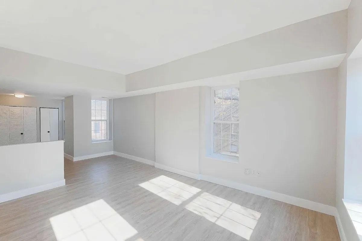 Bright, empty room with large windows allowing natural light to fill the space. The walls are painted a soft neutral color, and the flooring is light wood. There is a small partitioned area on the left. The overall atmosphere is airy and minimalist, suitable for various designs or purposes.