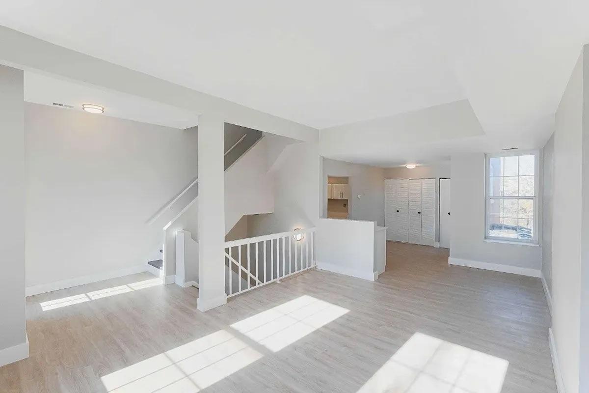 Interior of a bright, modern living space featuring light-colored walls and hardwood floors. The open layout includes a staircase on the left, a large window allowing natural light, and a doorway leading to another room. The area is well-lit and spacious, ideal for various furniture arrangements.