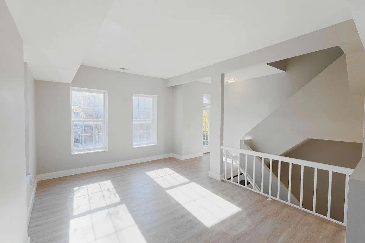 Spacious, bright room with large windows letting in natural light, light-colored walls, and light wood flooring. A staircase is visible in the corner, leading to another level, with a modern railing. The overall atmosphere is open and airy, perfect for various uses, such as a living area or office space.