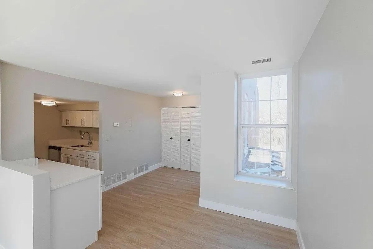Bright, empty interior of a modern apartment featuring a living area with light hardwood floors, a small kitchen visible in the background, and large windows providing natural light. The walls are painted in a light color, creating a spacious and airy atmosphere.