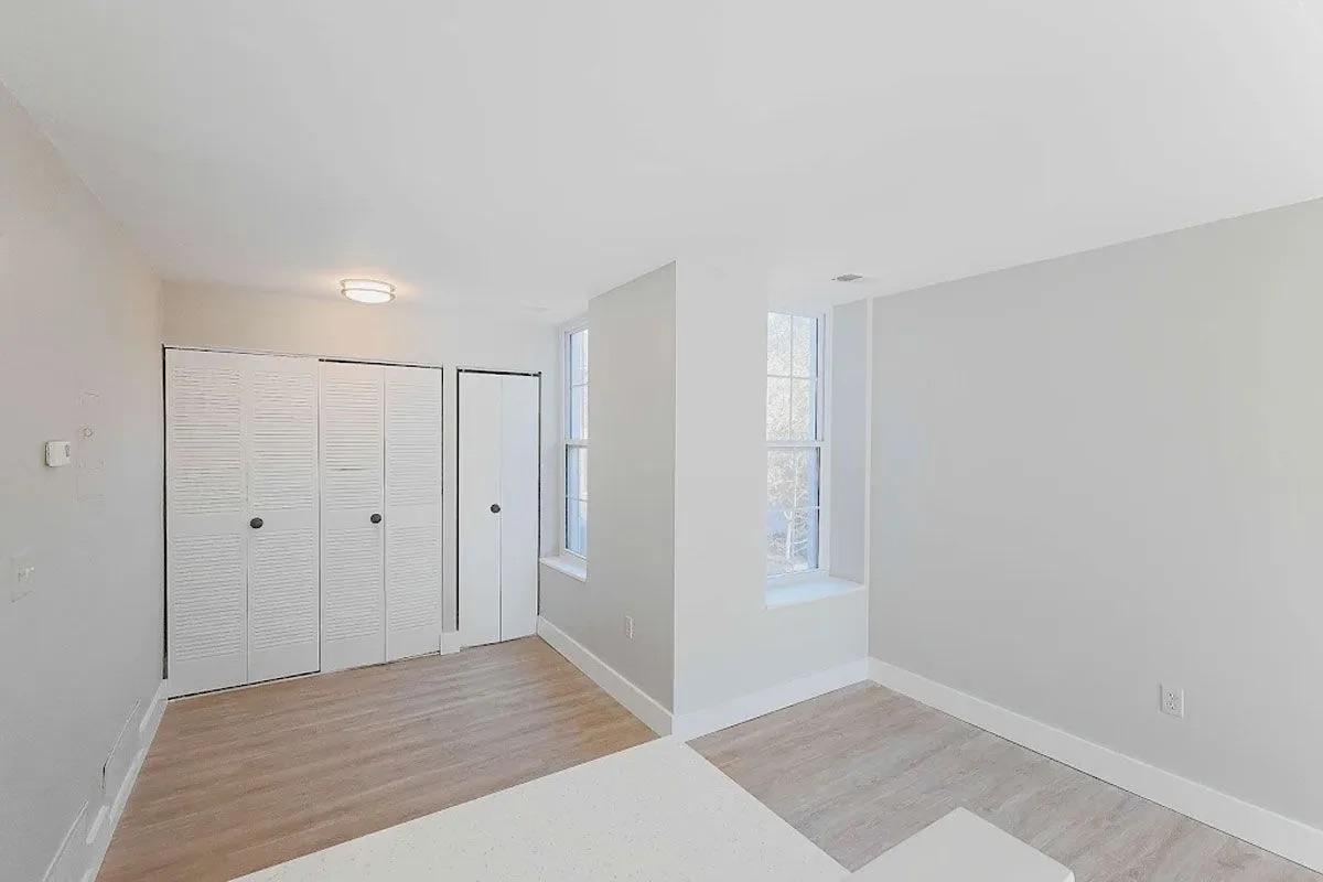 A bright, empty room featuring light-colored walls, a large window allowing natural light, and a laminate floor. There are built-in closets with sliding doors on one side, and a small section of the room is visible with white trim and a ceiling light fixture. The space has a clean and modern look.