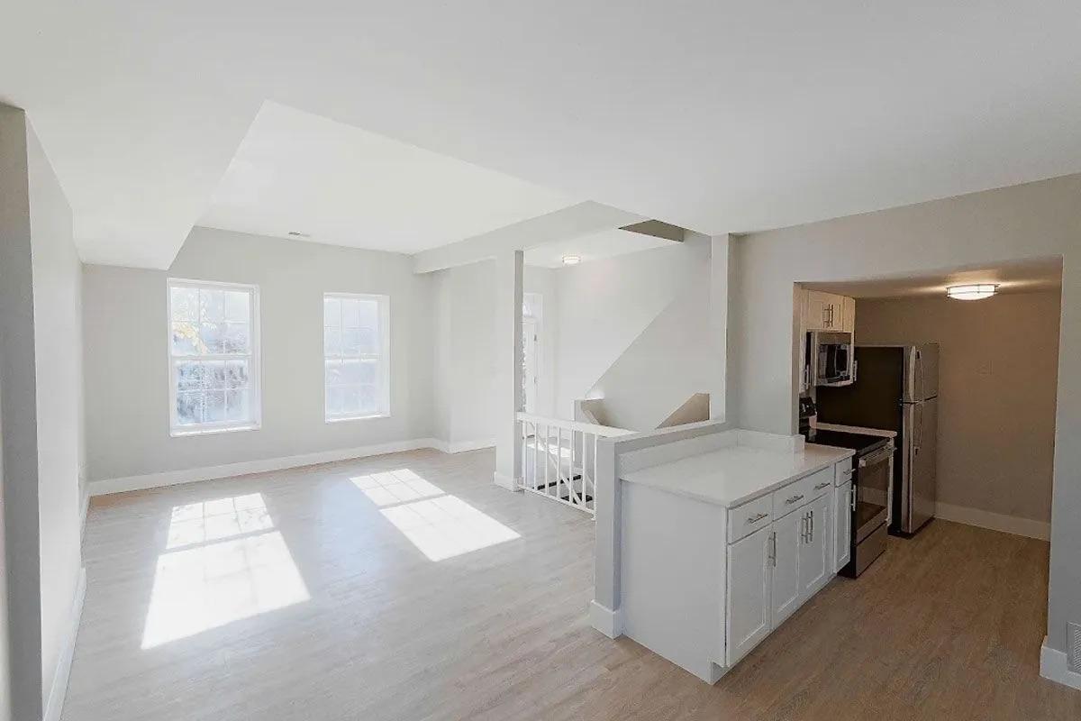 A spacious, modern living area with hardwood floors, large windows allowing ample natural light, and a staircase leading to an upper level. The open kitchen features white cabinetry and stainless steel appliances, creating a bright and inviting atmosphere.