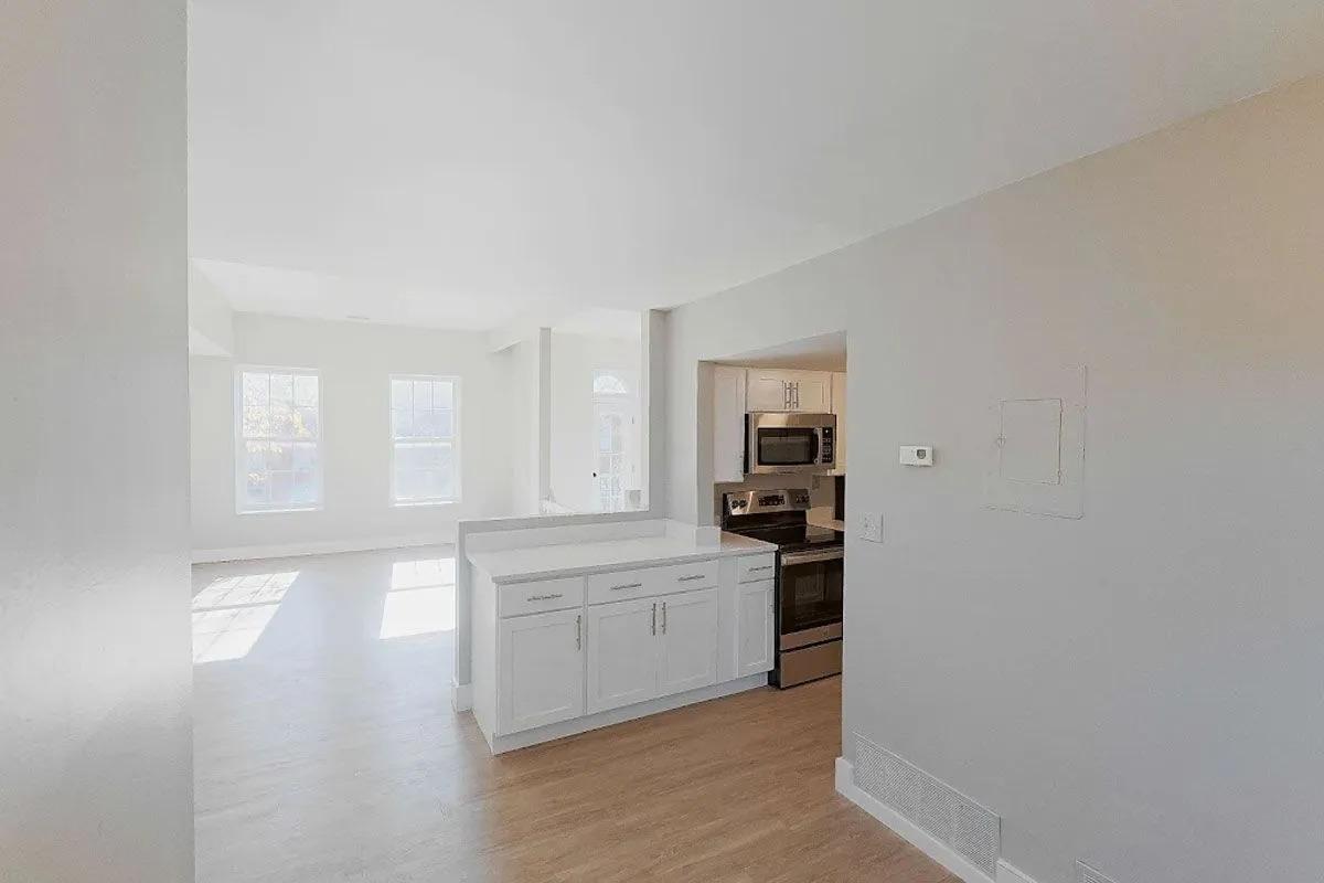 A bright, modern kitchen open to a spacious living area. The kitchen features white cabinetry and stainless steel appliances, including a microwave and oven. Large windows allow natural light to fill the space, creating an airy atmosphere with light-colored flooring and neutral walls.