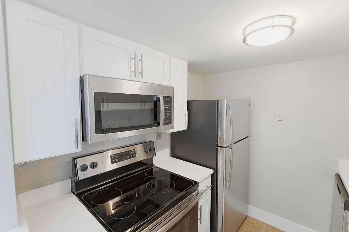 A modern kitchen featuring a stainless steel microwave above a stove, a refrigerator on the right, and white cabinetry. The countertop is light-colored, and there is a ceiling light providing bright illumination. The walls are painted in a neutral tone.