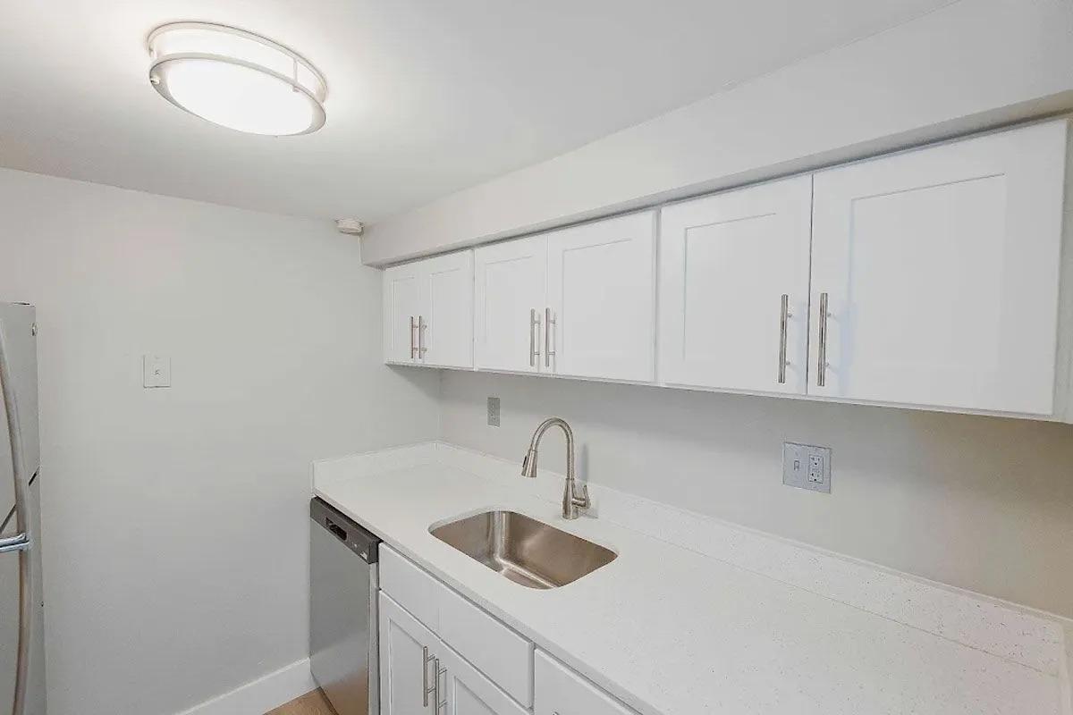 Modern kitchen with white cabinetry, a stainless steel sink, and a dishwasher. The countertop is light-colored and smooth, complementing the clean, minimalist design. A ceiling light illuminates the space, creating a bright and airy atmosphere.