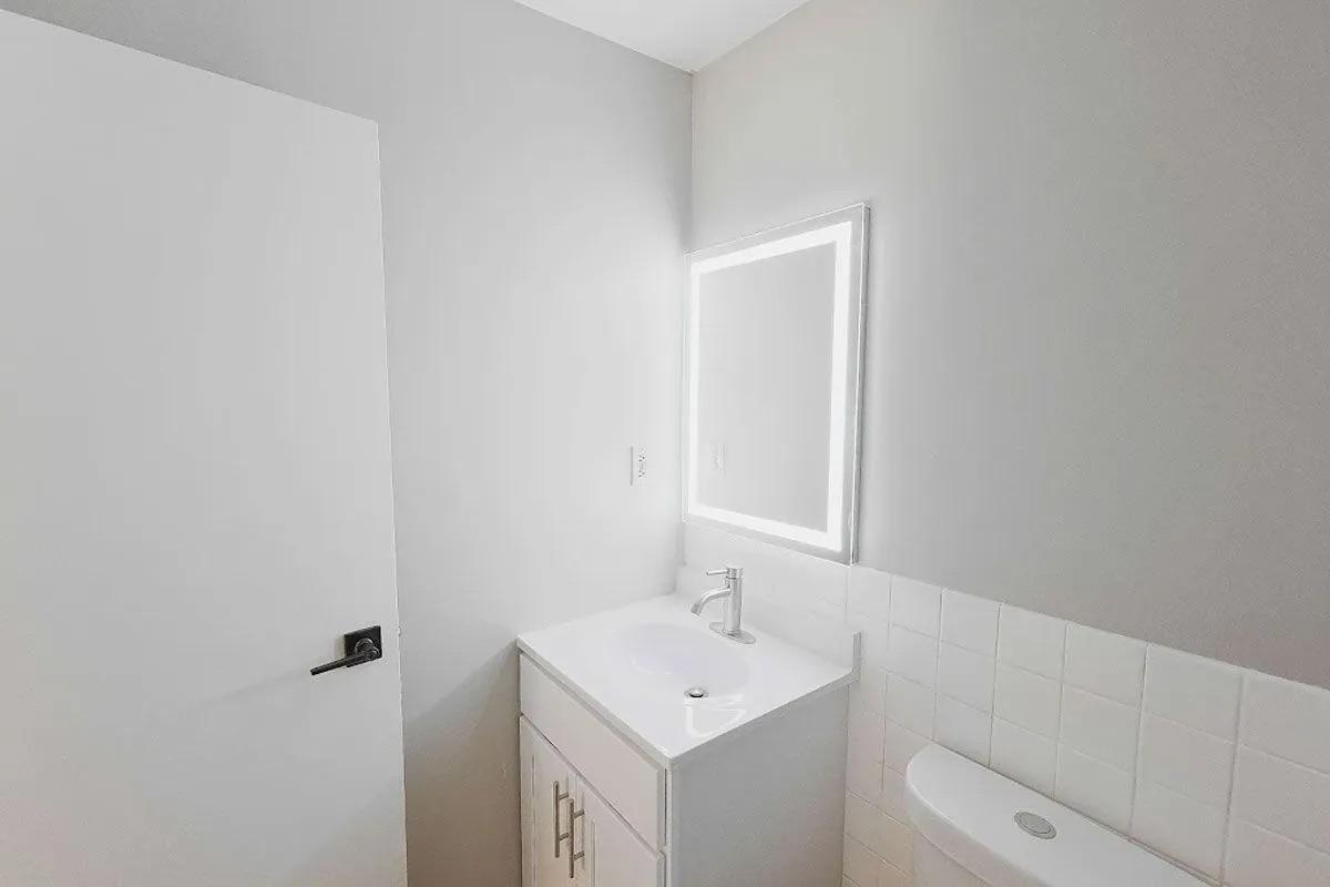A modern bathroom with light gray walls, featuring a white vanity with a sink, a mirror with built-in LED lighting, and a toilet against a white tiled wall. Natural light illuminates the space from an unseen window. The door is closed, adding to the minimalist aesthetic.