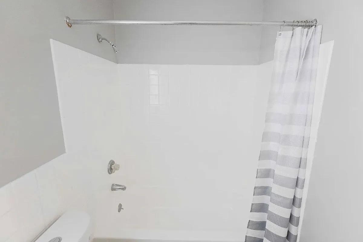 A clean and modern bathroom featuring a white tiled shower area with a silver shower rod. There is a striped shower curtain partially drawn, chrome fixtures, and a portion of a toilet visible at the bottom left. The walls are painted in a light gray color, creating a fresh and airy atmosphere.