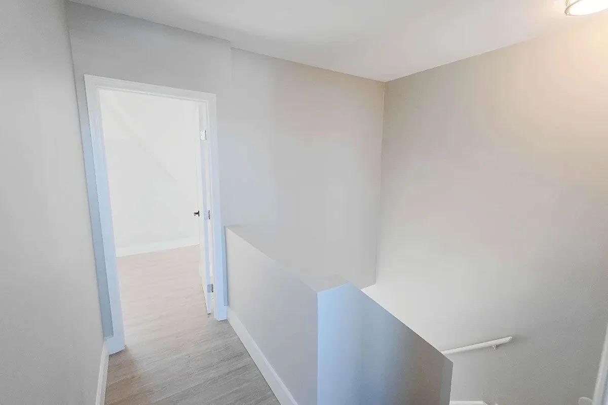 A well-lit, minimalistic staircase leading to a partially open room. The walls are painted a light color, creating a spacious feel. A door is visible at the end of the hall, and the lighting fixture casts a soft glow. The flooring appears to be light-colored wood, enhancing the clean aesthetic.