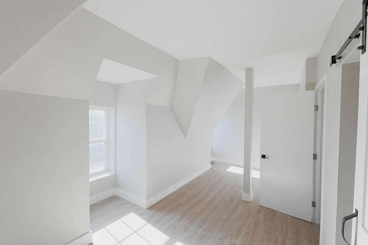 A brightly lit interior room featuring light-colored walls and a wooden floor. The space includes an angled ceiling, a large window allowing natural light, and a white door partially visible. The overall design is modern and minimalistic, with a sense of openness.