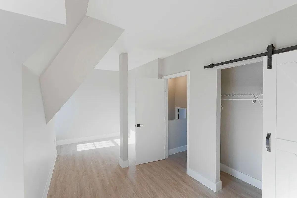 Interior view of a bright, modern room with light-colored walls and hardwood flooring. Features include a sloped ceiling, a closed white door, and an open closet with a hanging rod. Natural light enters through a nearby window, creating an airy atmosphere.