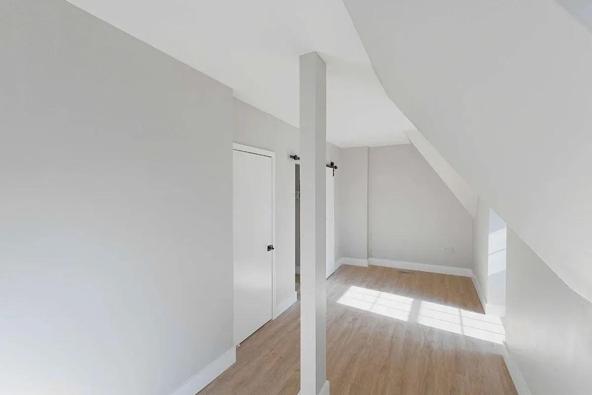 A spacious, light-filled room with light gray walls and a wooden floor. There is a white door on the left and a vertical white column in the center. Natural light streams in from a window, casting shadows on the floor. The room has a minimalist and modern feel.