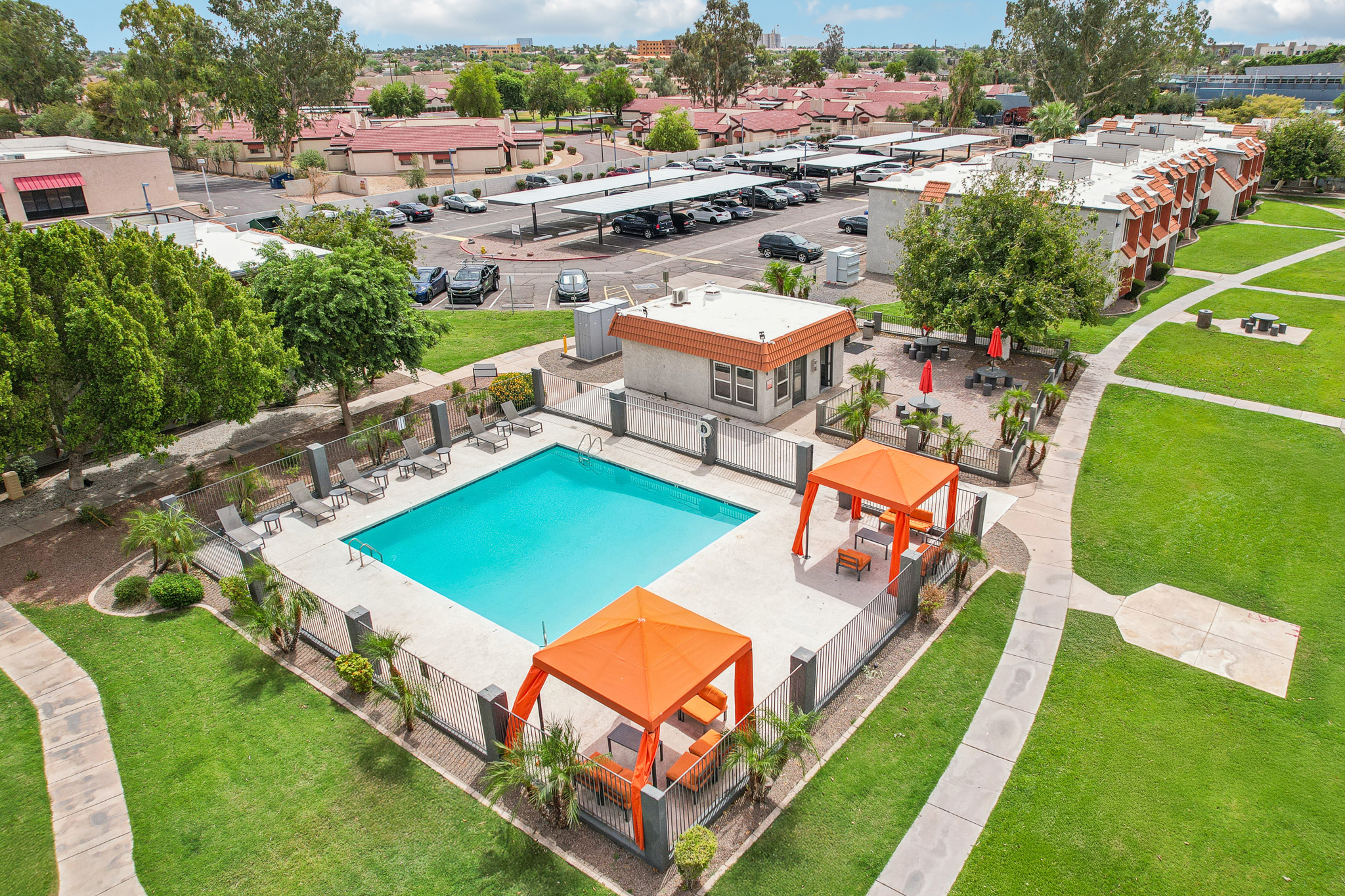 Aerial view of a community pool with bright orange cabanas and lounge chairs, surrounded by green grass and trees. Nearby, parking is visible with cars under shaded canopies, and residential buildings are in the background. The sky is partly cloudy, creating a vibrant atmosphere.