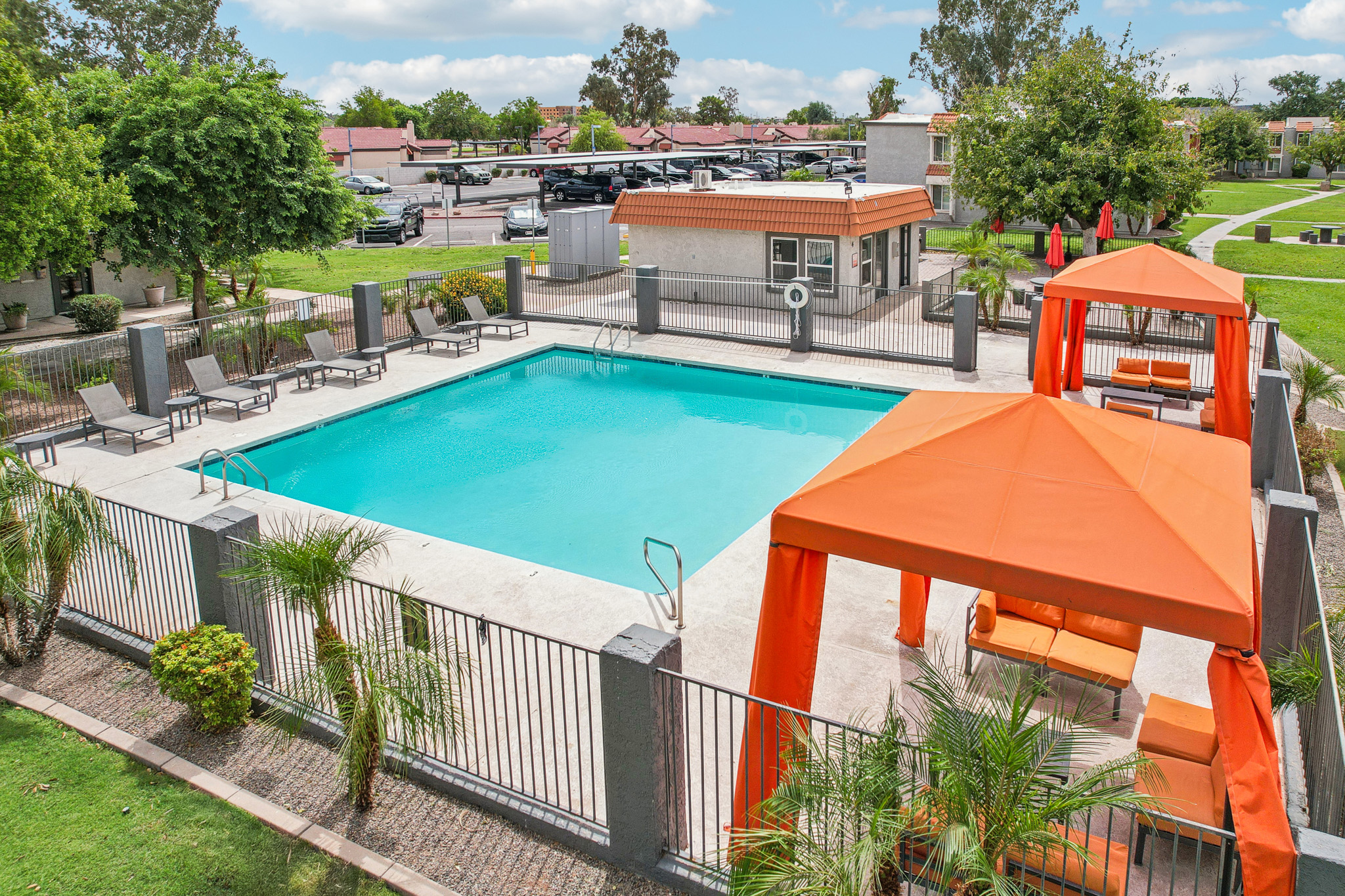 A spacious outdoor swimming pool surrounded by lounge chairs and orange cabanas. The pool area is enclosed by a fence and features well-maintained landscaping with palm trees. In the background, additional buildings and parking are visible under a clear blue sky.