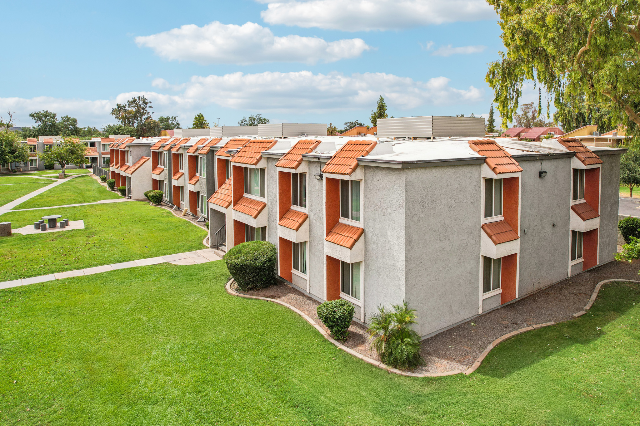 A view of a residential building with multiple units, featuring orange tiled roofs. The structure is surrounded by well-maintained green lawns, small bushes, and trees. The sky is partly cloudy, creating a pleasant atmosphere. Benches are visible in the grassy areas, suggesting a communal space.
