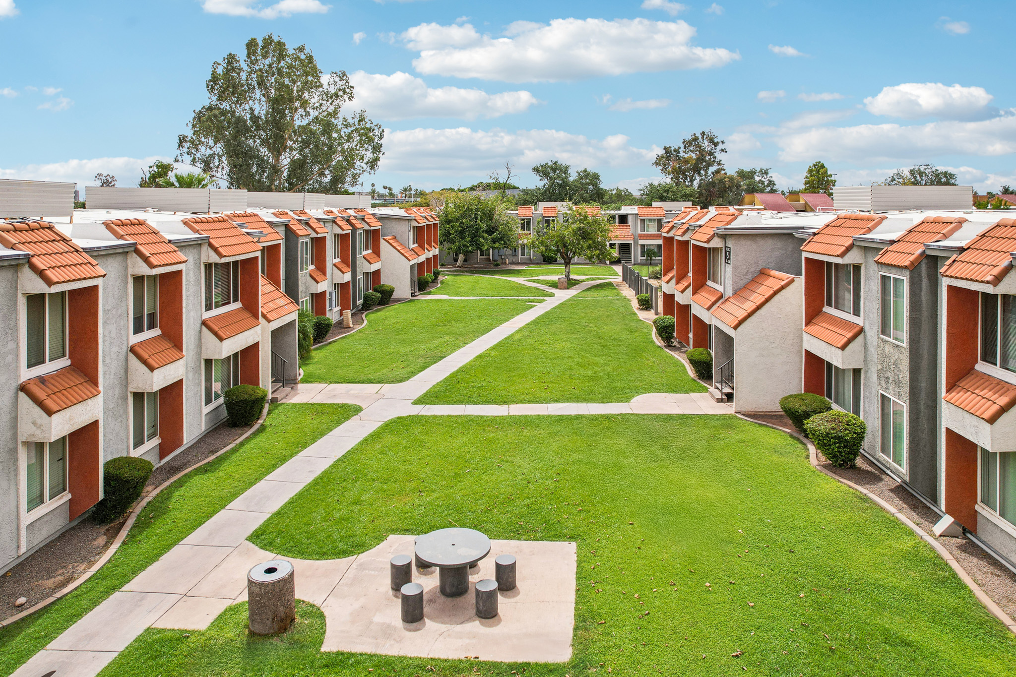 A view of a landscaped courtyard in a residential complex, featuring several two-story buildings with orange and gray exteriors. A grassy area separates the buildings, with a path running through it. In the center, there is a circular table with benches, surrounded by well-maintained shrubs and trees, under a partly cloudy sky.